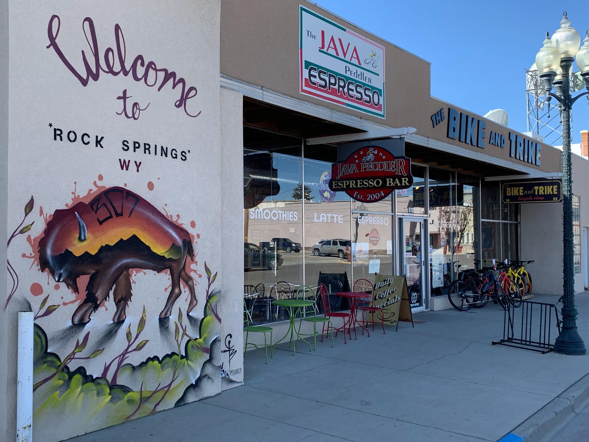 A storefront in Rock Springs, Wyoming, featuring a colorful mural of a bison and the text "Welcome to Rock Springs WY 307." The building houses two businesses: "The Java Peddler Espresso" and "The Bike and Trike." Outdoor seating with brightly colored chairs is visible, along with bicycles parked outside. The scene is set on a clear, sunny day.
