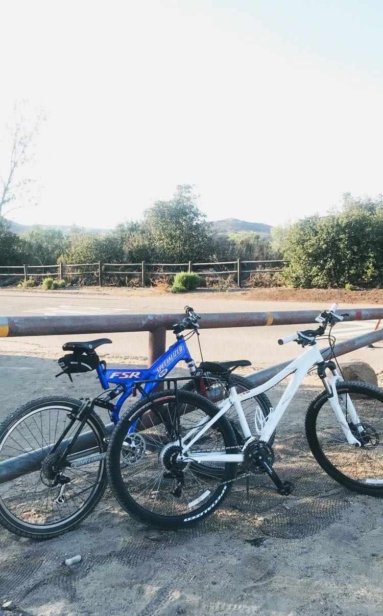 Specialized Myka 26: Two mountain bikes, one blue and one white, are leaning against a railing in a natural outdoor setting. The background features green trees and an unpaved road, with hills visible in the distance. The bikes are positioned close to each other on a dirt surface.