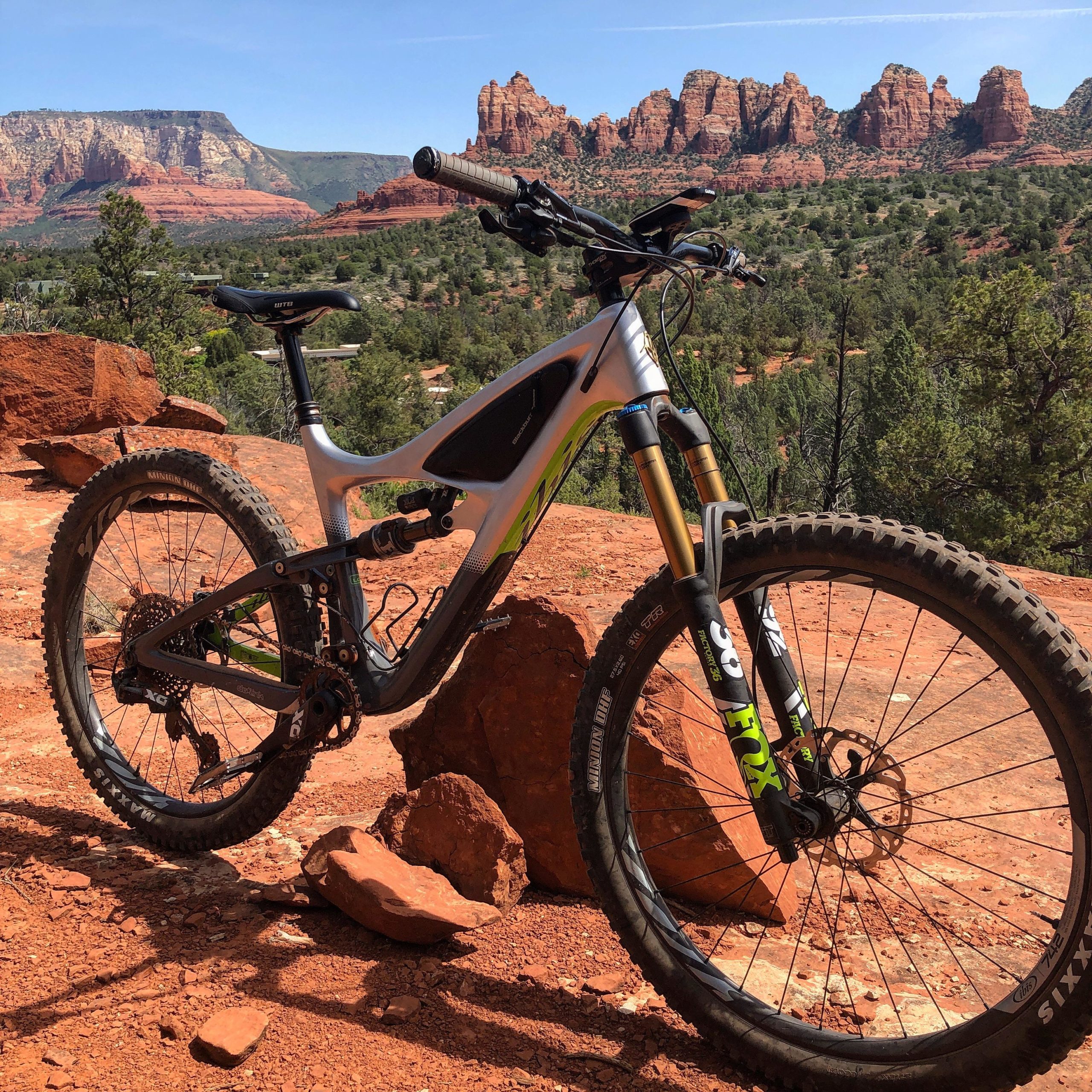 A mountain bike positioned on a rocky terrain, with a backdrop of red rock formations and green pine trees under a clear blue sky. Pig Tail mountain bike trail.