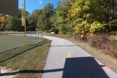 A curved paved pathway for walking or biking, bordered by a chain-link fence and surrounded by trees with autumn foliage. A sign is visible in the distance, indicating the path leads toward a recreational area. DuPage River Sports Complex mountain bike trail.