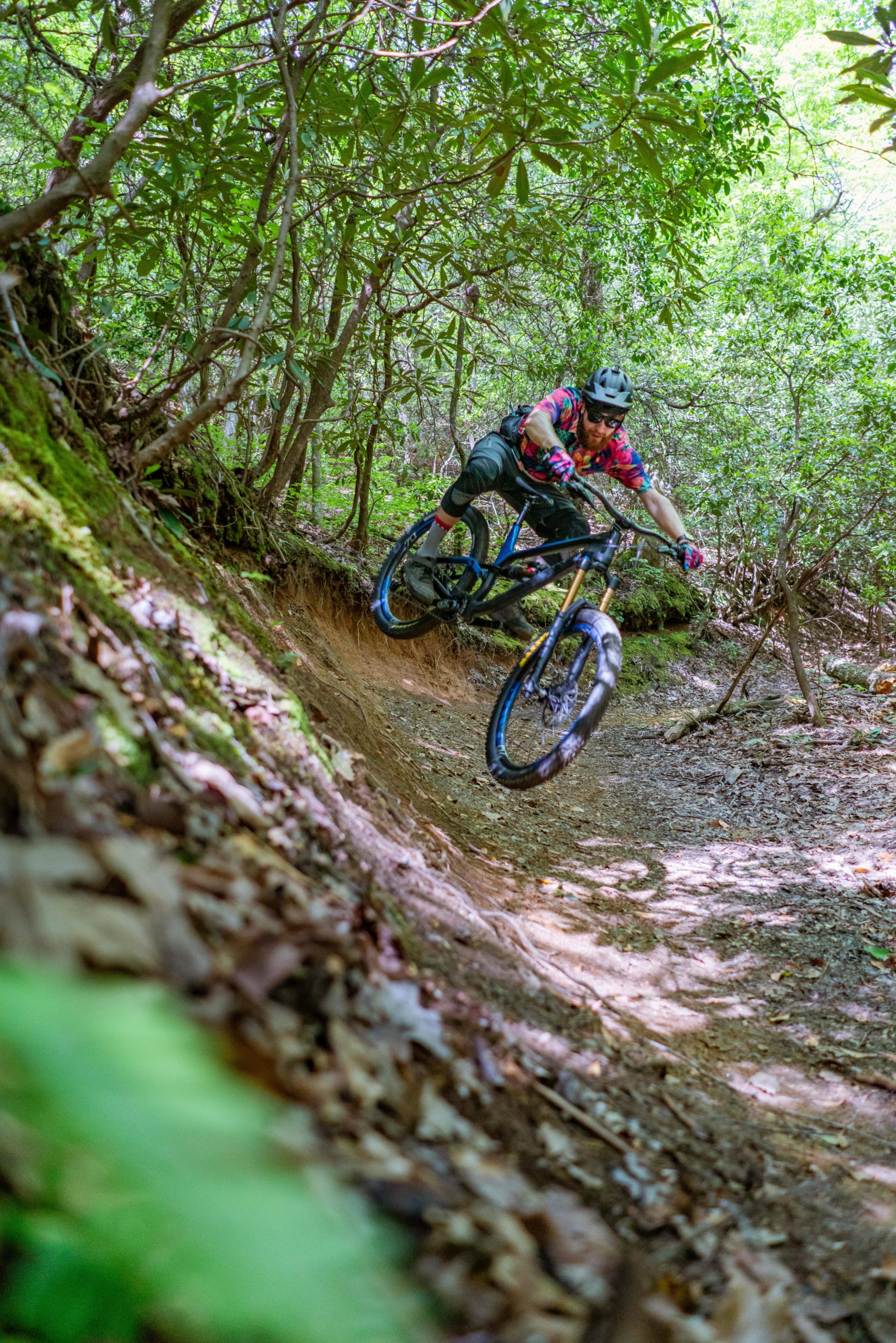 A mountain biker navigating a dirt trail in a lush, green forest. The cyclist is wearing a helmet and colorful biking gear, leaning into a turn as they maneuver a challenging bend on the track. Sunlight filters through the trees, creating dappled shadows on the ground. Bracken Preserve mountain bike trail.