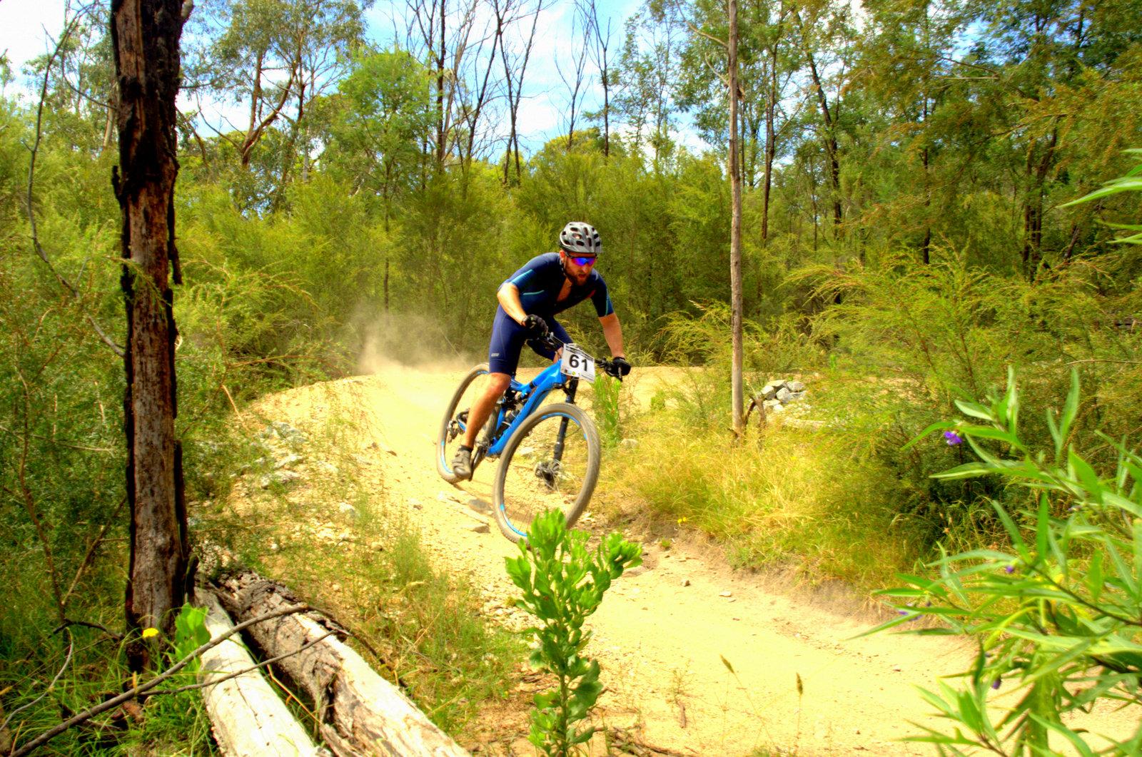 A mountain biker in a blue and black outfit navigates a dirt trail surrounded by greenery, kicking up dust as he speeds around a curve. The scene captures the excitement of off-road cycling in a natural setting, with tall trees and foliage in the background. Lysterfield Mountain Bike Area mountain bike trail.