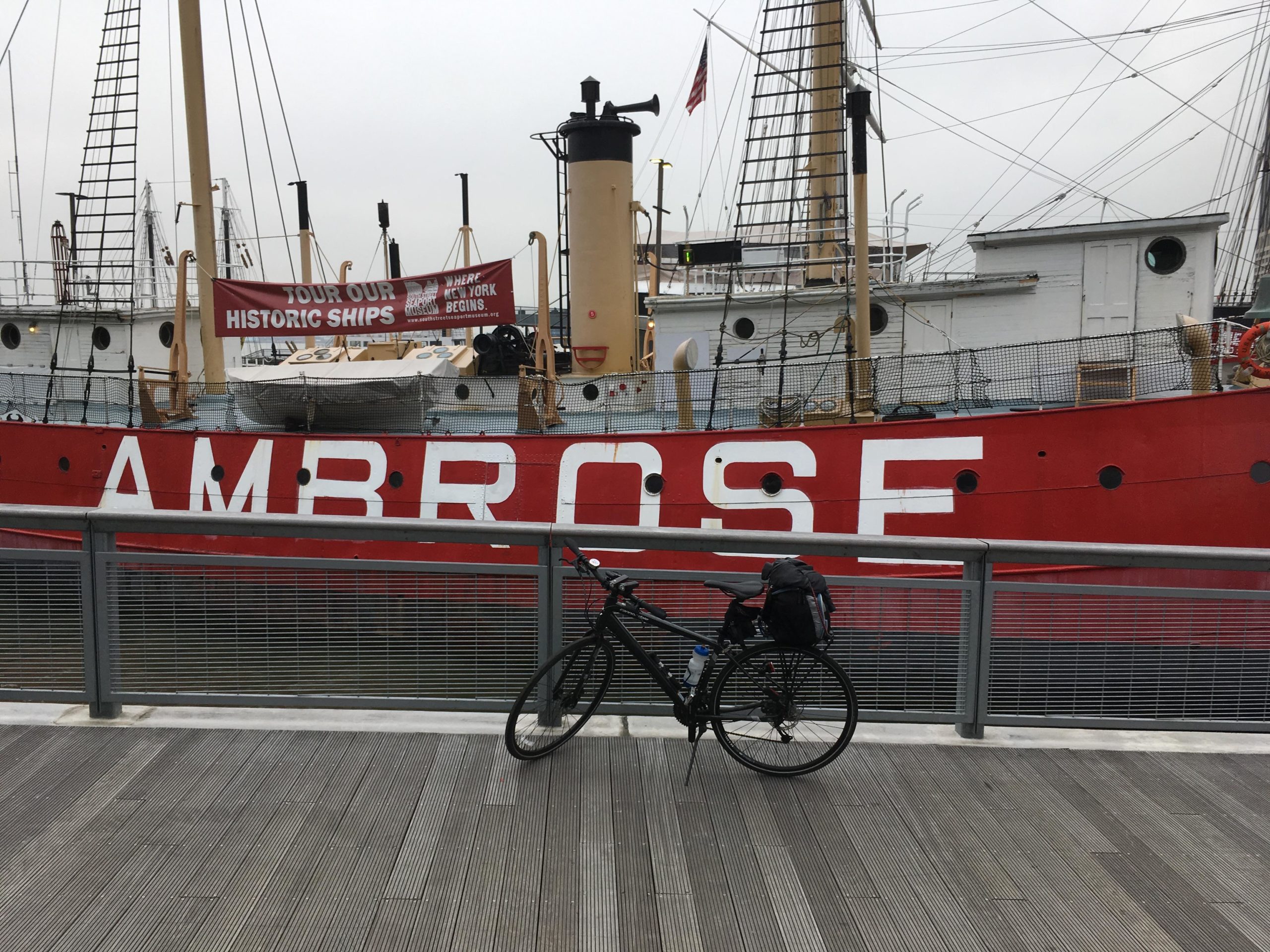 A bike is parked on a wooden dock in front of the historic ship Ambrose, which features prominent red and white lettering. A banner above the ship invites visitors to tour historic ships. The scene is set on a cloudy day, with masts of other boats visible in the background. East Side Green way 34th st to the Staten Island Ferry mountain bike trail.