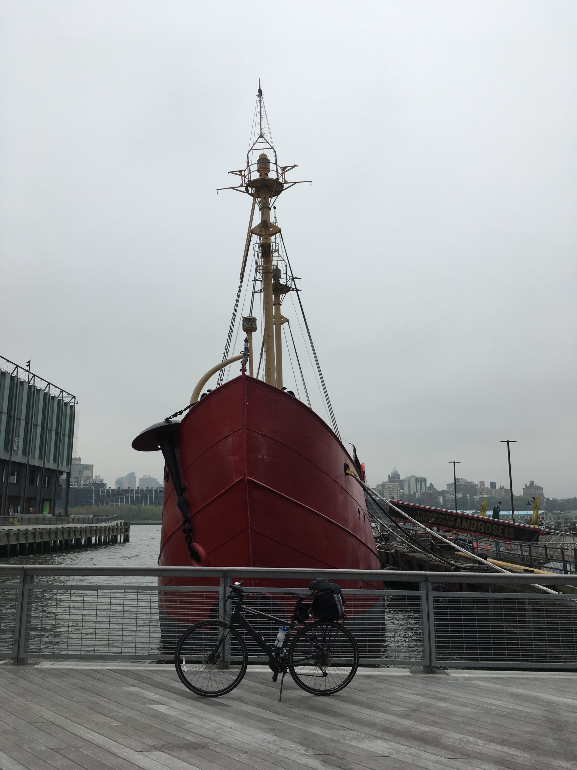 A large red ship docked at a pier, with its mast prominently displayed against a gray sky. In the foreground, a black bicycle is parked on a wooden boardwalk by the water, adding a sense of scale to the scene. The background features buildings and a distant skyline. East Side Green way 34th st to the Staten Island Ferry mountain bike trail.