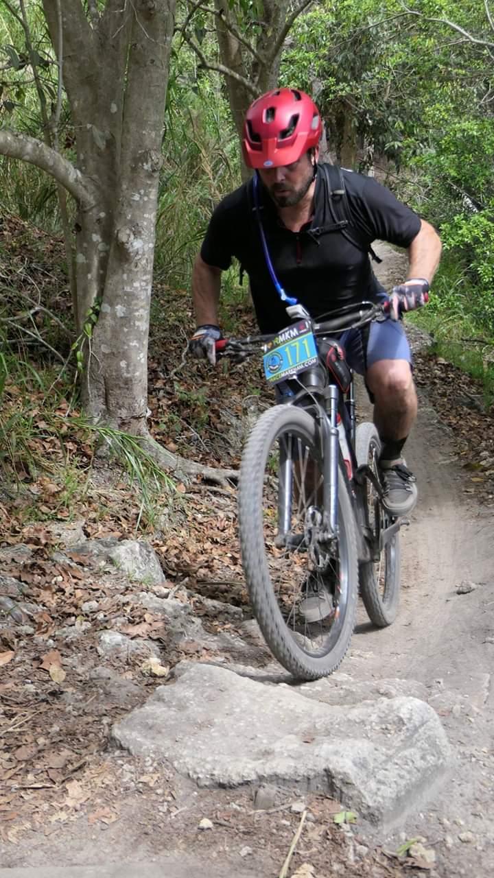 A mountain biker in a red helmet rides over a rocky trail in a lush, green forest. The rider is focused on navigating the terrain, with trees and foliage in the background. Markham Park mountain bike trail.