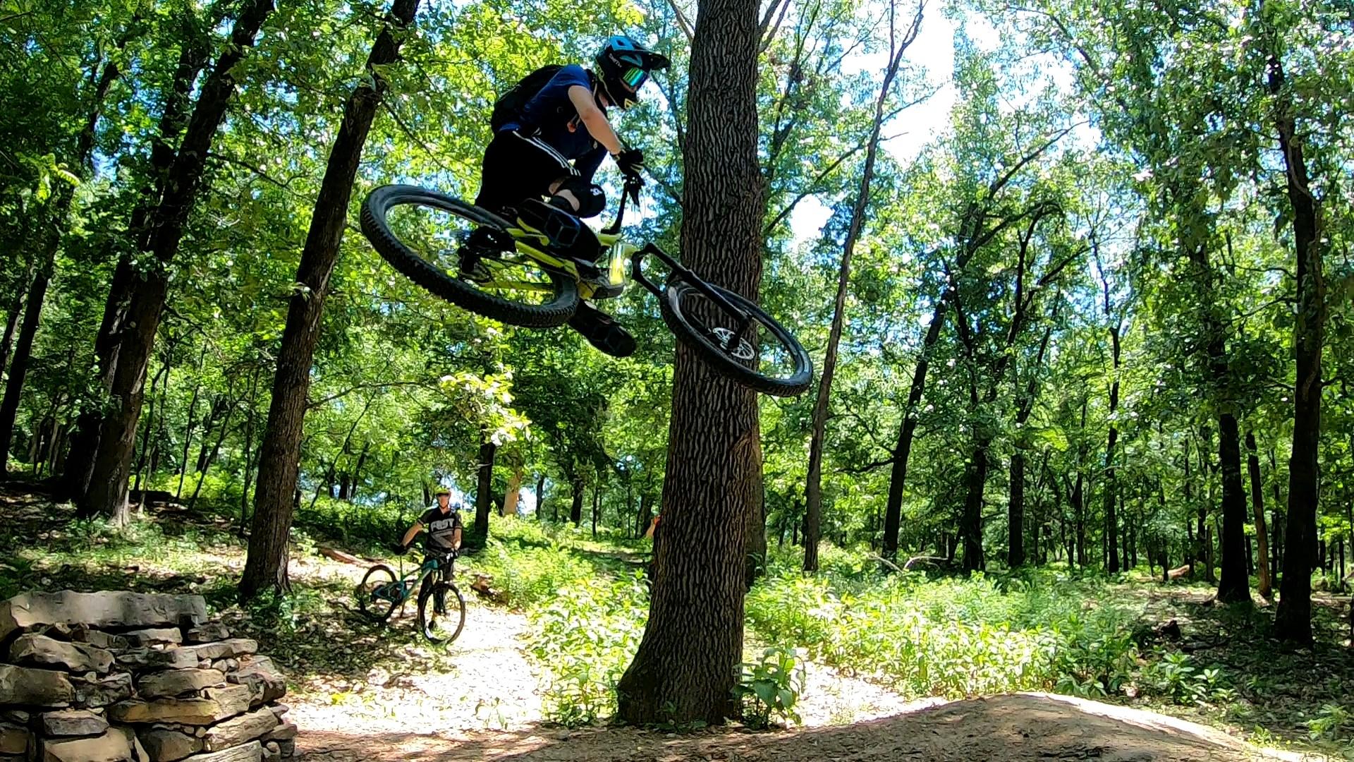 A mountain biker performs a jump in a forested area, showcasing aerial skills while another rider waits nearby on the trail. Towering trees and lush greenery surround the scene, illuminated by bright sunlight. Slaughter Pen Trail mountain bike trail.