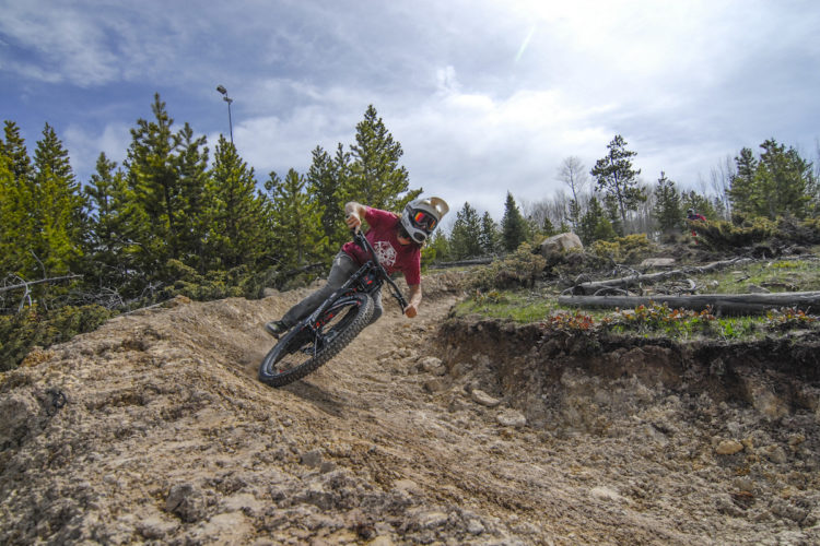 A person riding a mountain bike on a dirt trail, surrounded by trees and under a cloudy sky. The rider is leaning into a turn, wearing a helmet and protective gear. The terrain is uneven with rocks and dirt, typical of a biking course.
