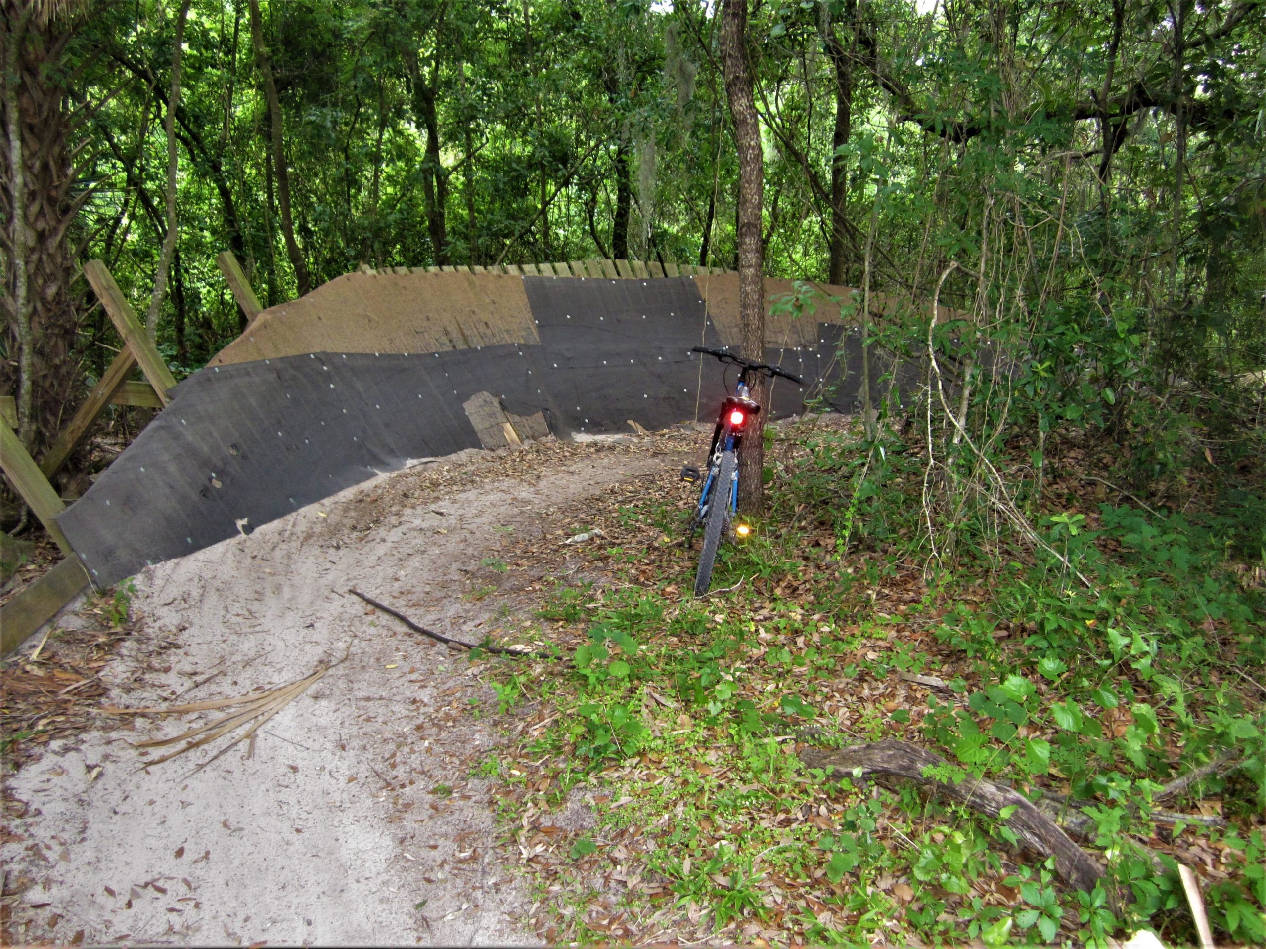 A mountain bike is parked near a dirt trail that curves around a wooden structure, surrounded by lush green foliage in a wooded area. The trail is sandy, and the structure appears to be an embankment designed for biking. Loyce E. Harpe Park mountain bike trail.