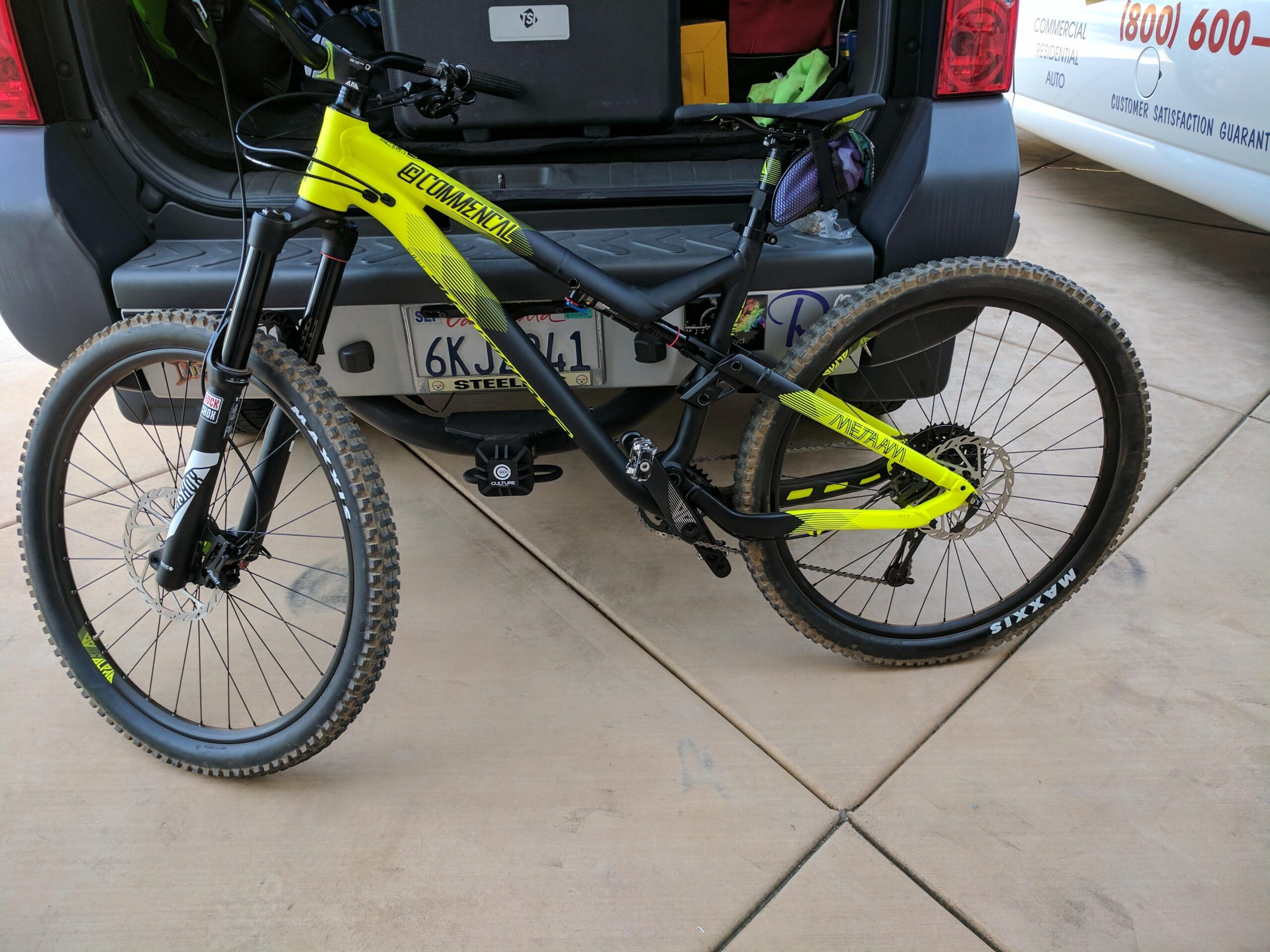 Commencal Meta AM V4: A brightly colored mountain bike with a distinctive yellow and black frame is parked beside an open vehicle. The bike features front suspension, wide tires with visible tread, and a disc brake. The vehicle's interior is partially visible, showcasing storage space. The surrounding surface is a concrete driveway.