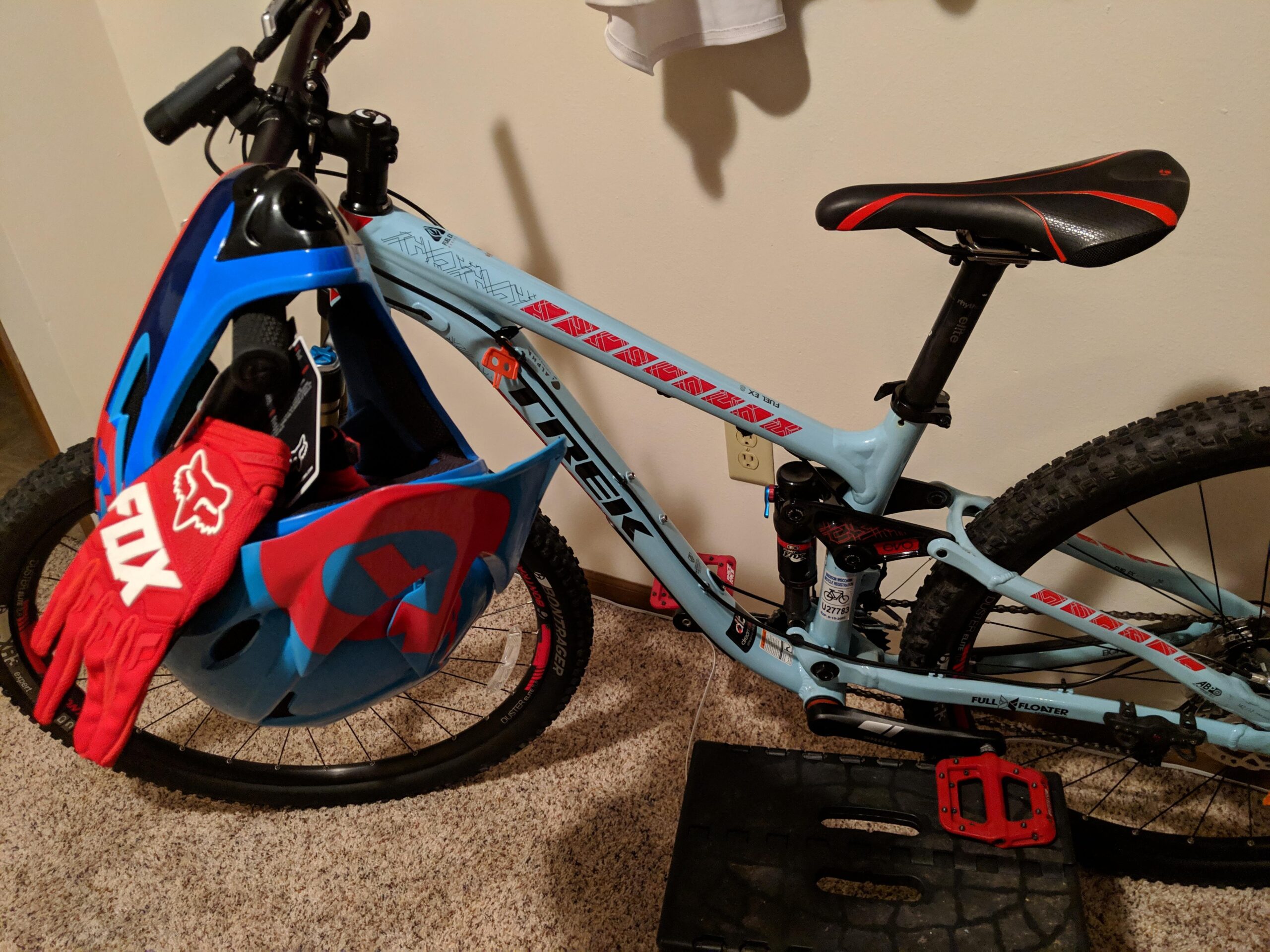 Trek Fuel EX 8 29: A blue and red mountain bike is positioned against a wall, featuring a helmet and red gloves resting on the front. The bike has chunky tires, a prominent seat, and a visible suspension system. A black platform is located beneath the bike, while the background shows a neutral-colored wall.