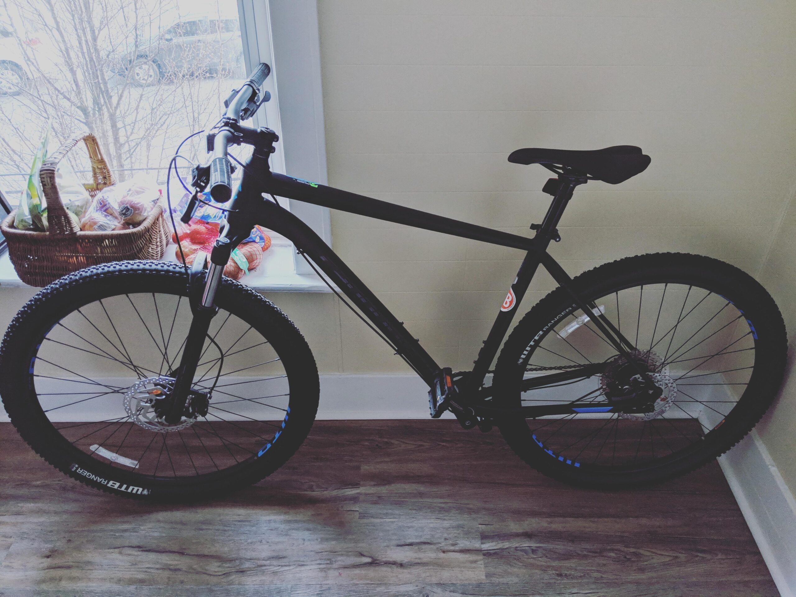Cannondale Trail 29er 5: A black mountain bike leaning against a wall, with a visible front wheel and handlebars. In the background, there is a window with a basket of assorted food items on the windowsill, including bread and other groceries. The flooring is wooden, and there is natural light coming through the window.