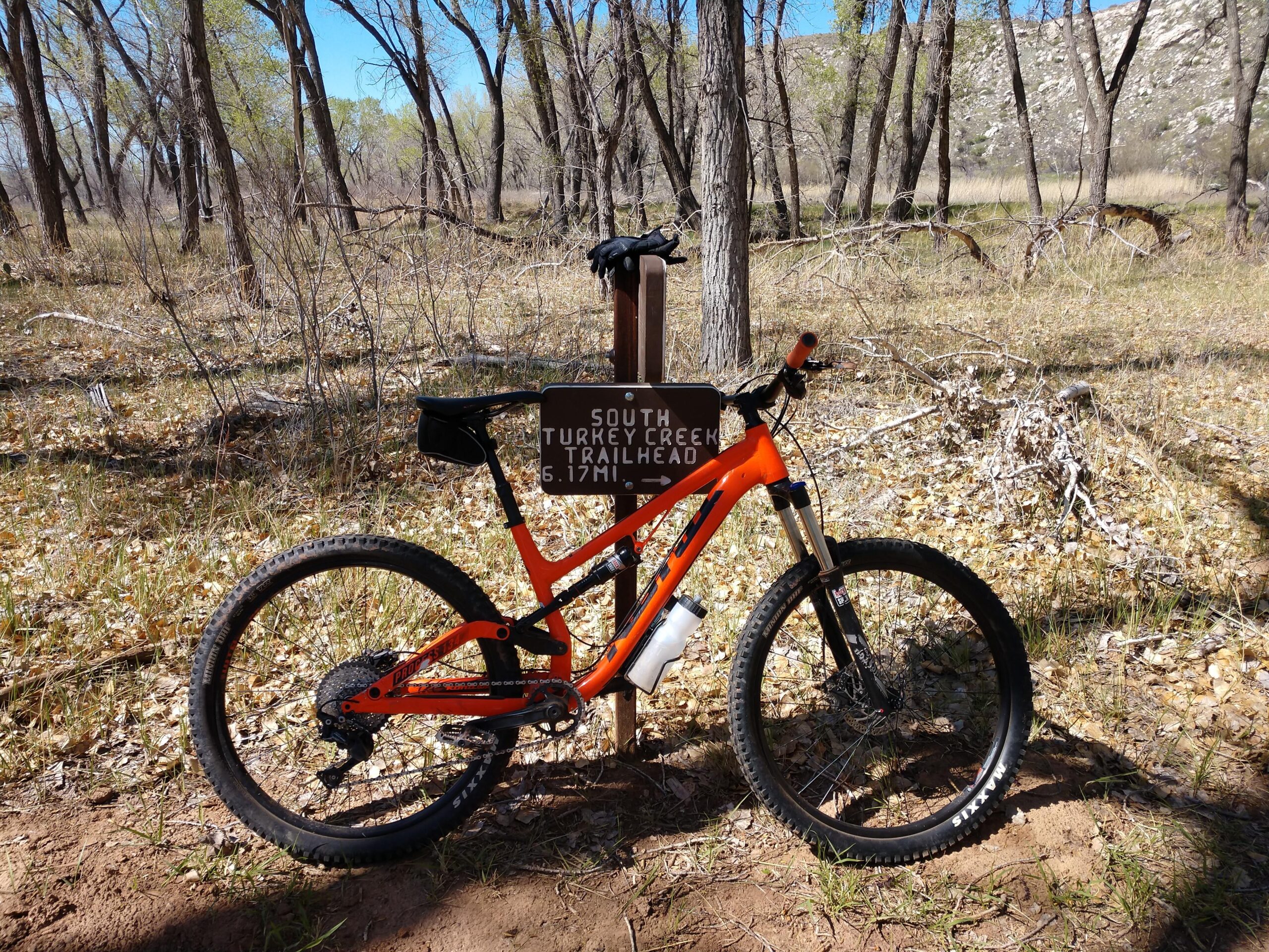 Kona Process 134: A mountain bike leaning against a trail sign that reads "South Turkey Creek Trailhead 6.17 mi." The bike is bright orange with black accents and is positioned on a dirt path surrounded by trees and sparse foliage, under a clear blue sky.