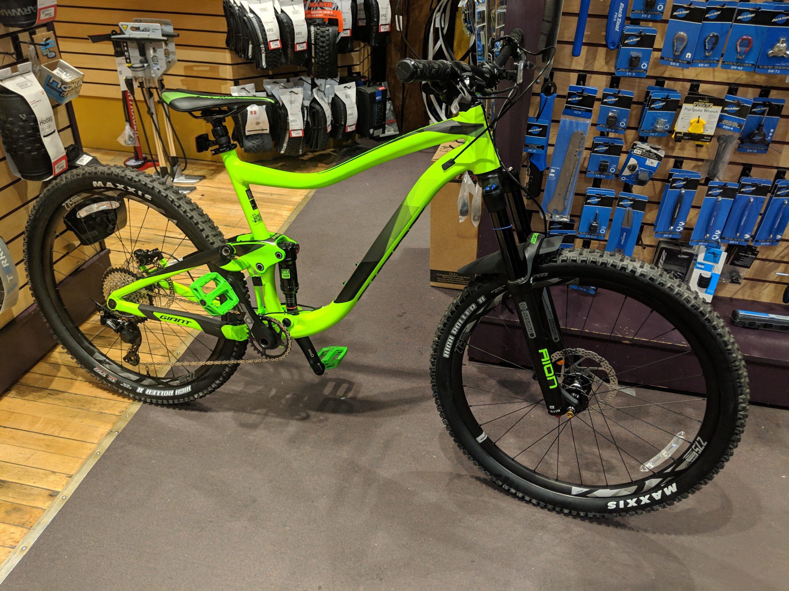 Giant Trance 27.5 3: A bright green mountain bike is displayed in a shop, showcasing its sleek design and components. The bike features large tires, a sturdy frame, and advanced suspension. In the background, various bike accessories and tools are organized on wall-mounted displays. The floor is covered with a dark carpet, and the overall atmosphere suggests a well-equipped cycling shop.