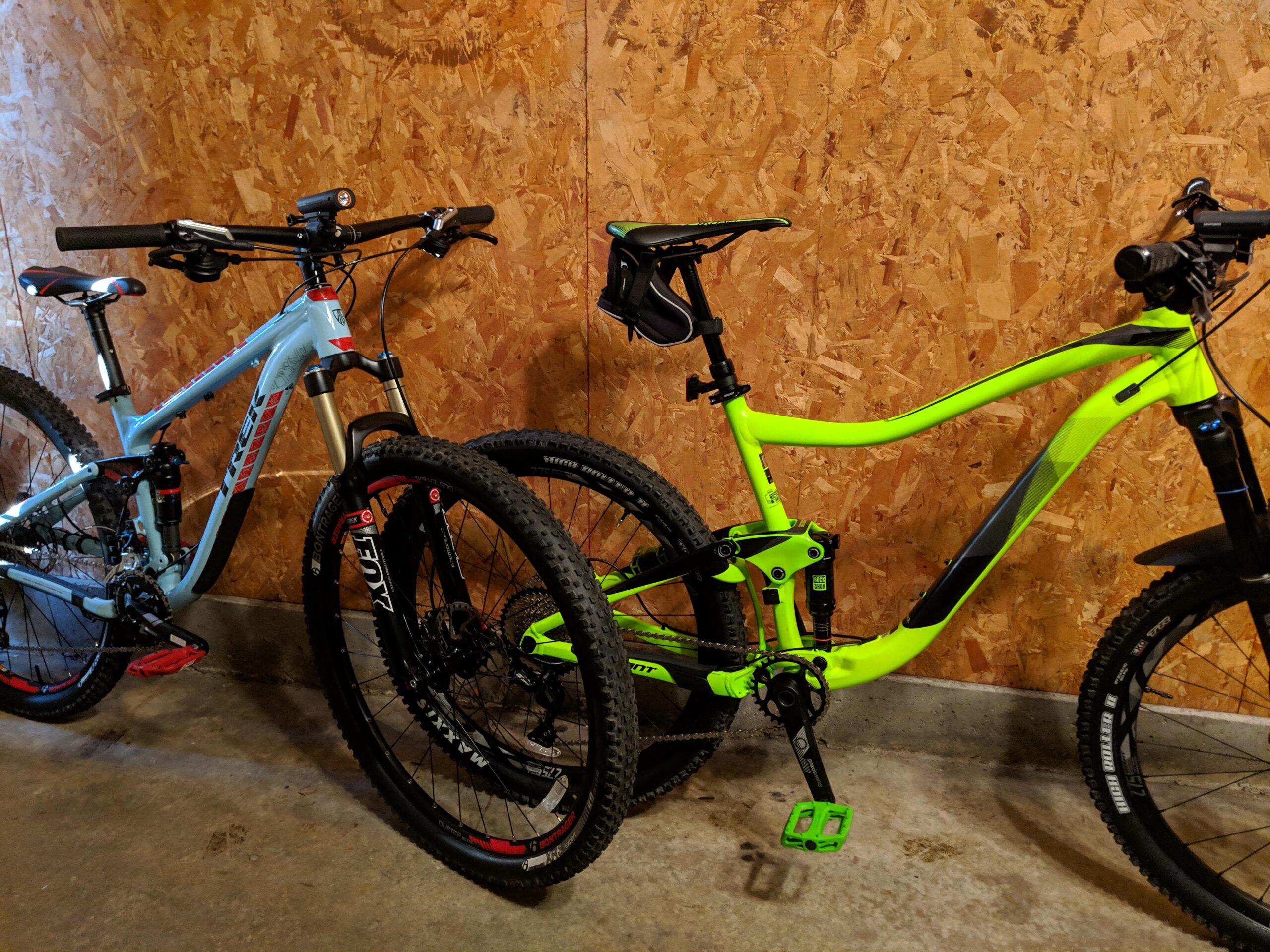 Giant Trance 27.5 3: Two mountain bikes are parked side by side against an orange wooden wall. The bike on the left is light blue with red accents and features a sturdy frame and wide tires. The bike on the right is bright green with black details and has a sleek design. Both bikes have mountain bike tires and are ready for outdoor adventures.