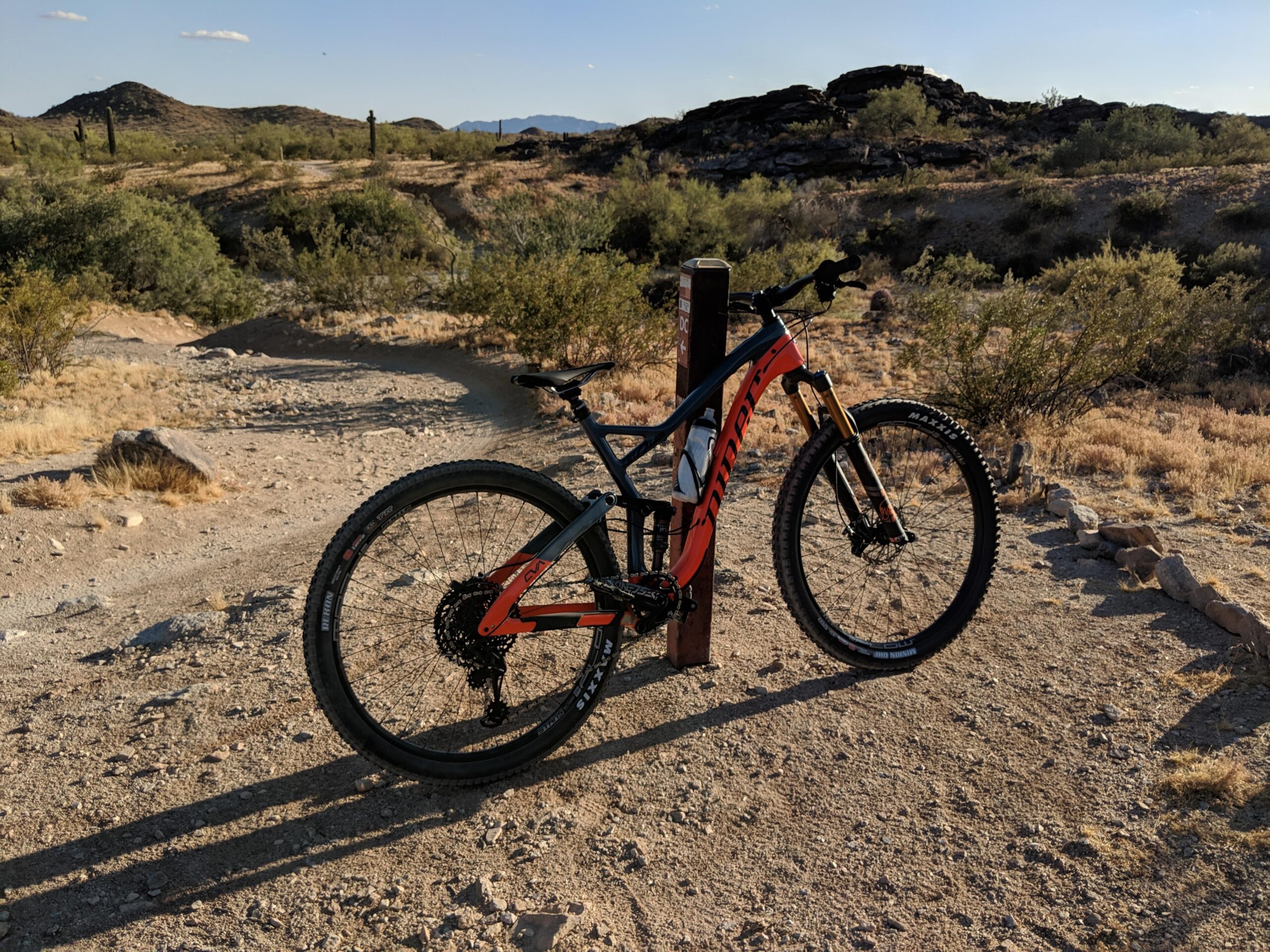 Niner Jet 9 RDO: A mountain bike is leaning against a wooden post on a dirt trail surrounded by desert vegetation and rocky hills in the background. The scene is bathed in warm sunlight, highlighting the bike's orange and black design.