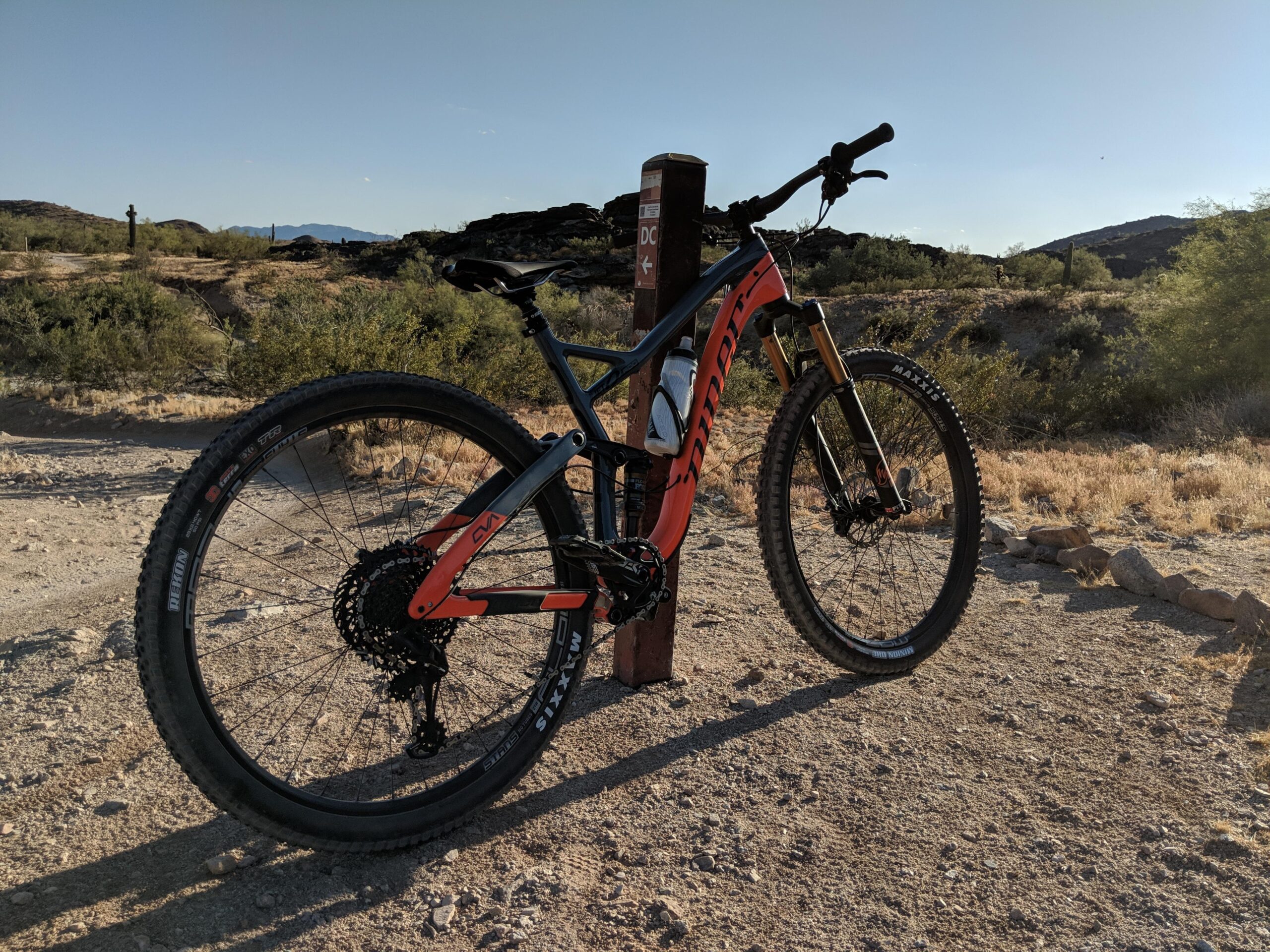 Niner Jet 9 RDO: A close-up view of a mountain bike with an orange and black frame, parked on a dirt path in a desert landscape. In the background, there are cacti and rocky hills under a clear blue sky. A trail sign is visible near the bike.