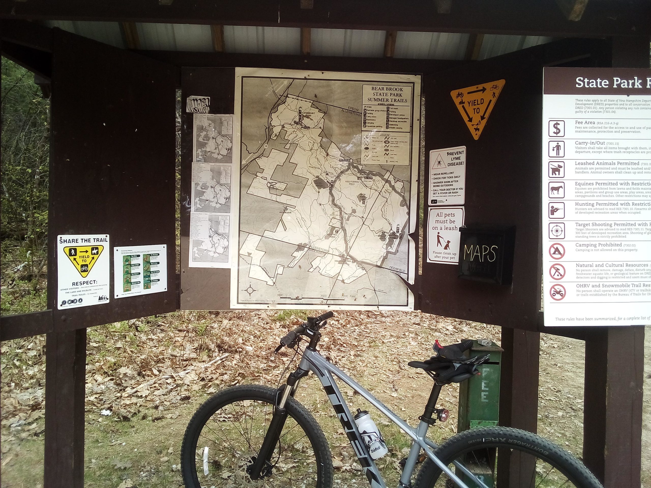 A trail map station at Bear Brook State Park featuring a large map of the park, informational signs about trail usage and safety, and a mountain bike parked beside the structure. The station includes notices for sharing the trail, rules for pets, and various park regulations. Bear Brook mountain bike trail.