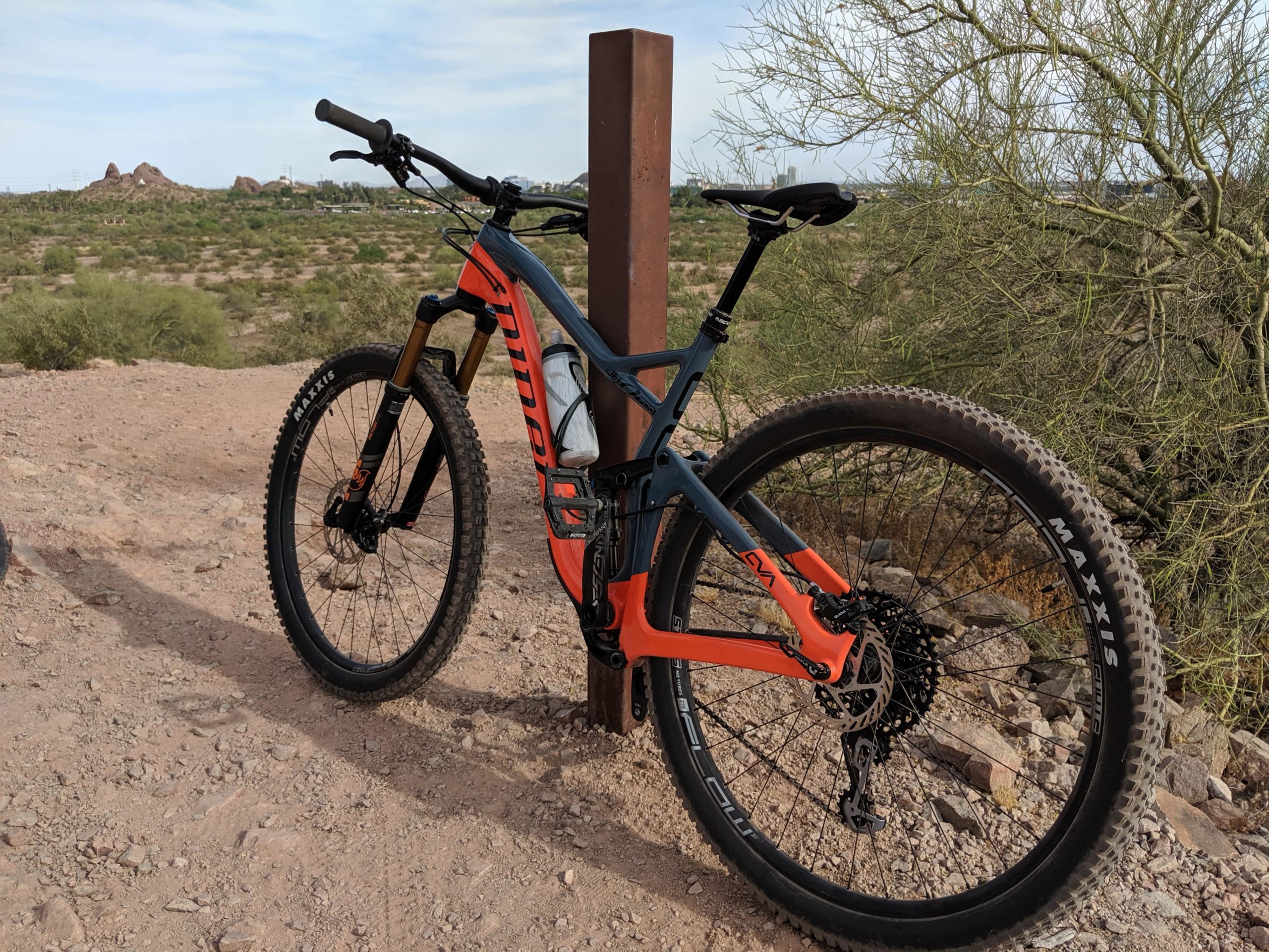 A mountain bike with an orange and black frame is parked near a signpost on a dirt trail surrounded by sparse vegetation and rocky terrain. In the background, distant hills are visible under a partly cloudy sky. Papago Park Area mountain bike trail.