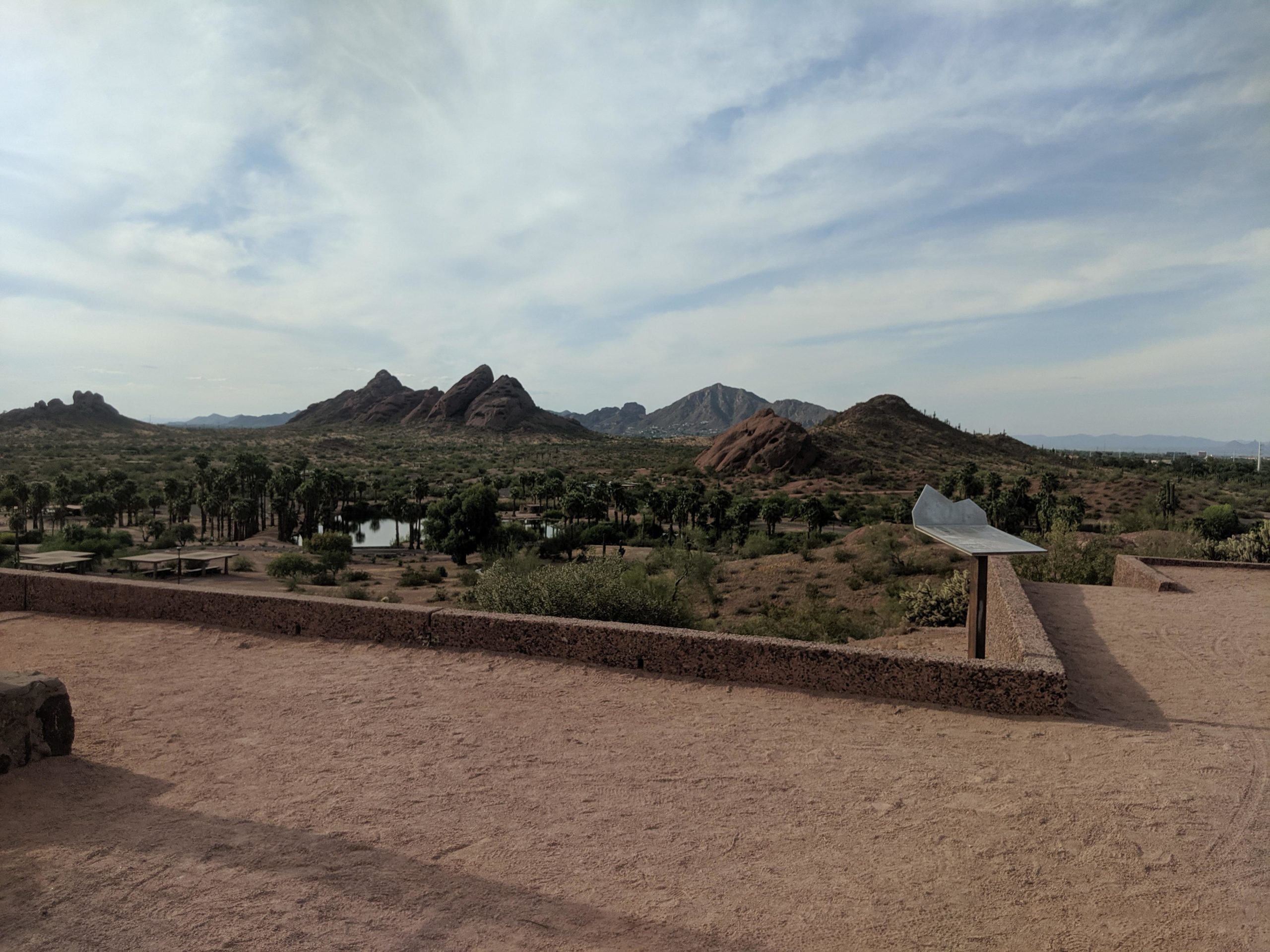 A scenic view of rocky mountains under a cloudy sky, with a desert landscape featuring sparse vegetation and a small pond in the foreground. There is a stone wall in the lower part of the image, and a metal sculpture on a pedestal. The area appears arid with gentle rolling hills in the distance. Papago Park Area mountain bike trail.