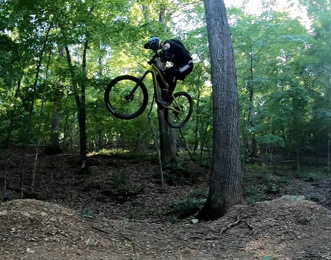 A cyclist performing a jump on a mountain bike in a forested area, surrounded by trees and greenery. The rider is airborne above a pile of dirt, with sunlight filtering through the leaves. Lake Leatherwood Gravity Project mountain bike trail.