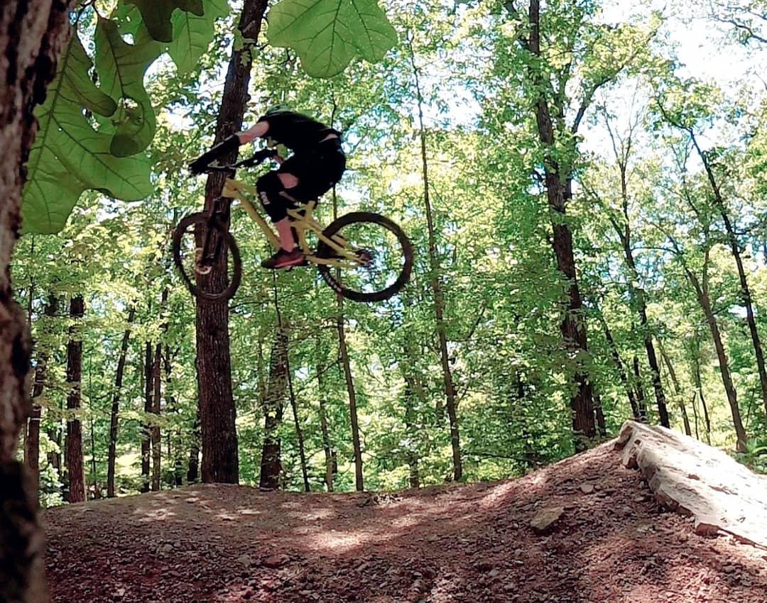 A person performing a mid-air jump on a mountain bike above a dirt ramp, surrounded by lush green trees in a forested area. Slaughter Pen Trail mountain bike trail.