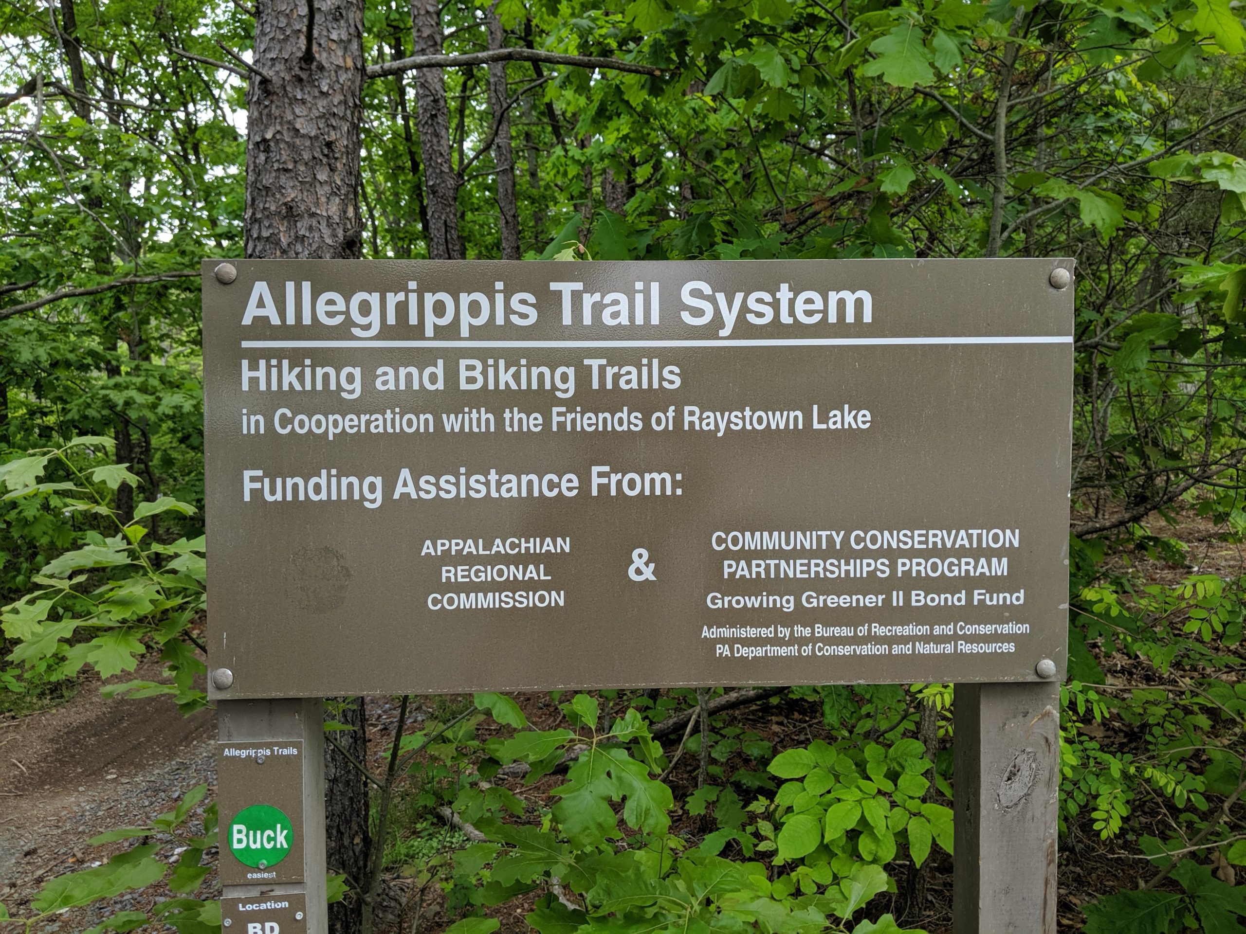 Sign for the Allegrippis Trail System, displaying information about hiking and biking trails in cooperation with the Friends of Raystown Lake. The sign notes funding assistance from the Appalachian Regional Commission and the Community Conservation Partnerships Program. Surrounding greenery and trees are visible in the background. Allegrippis Trails mountain bike trail.