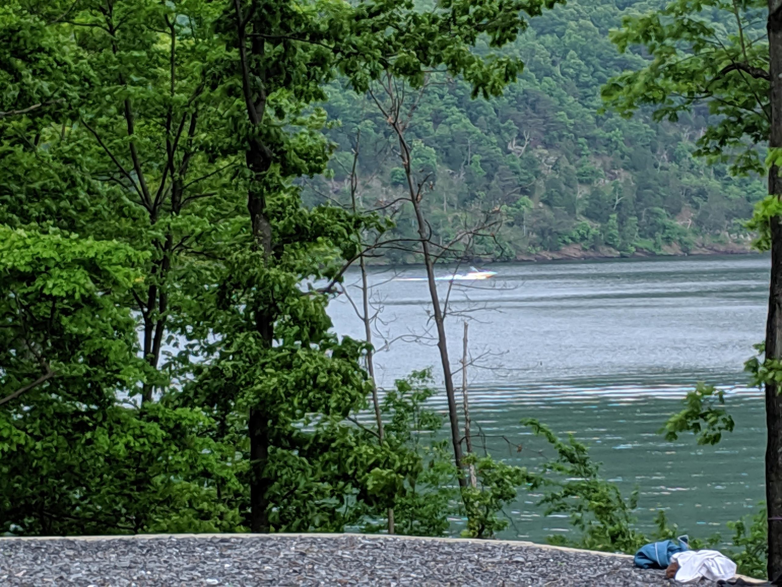 A serene view of a lake surrounded by lush green trees, with a small boat visible on the water in the distance. The foreground features some gravel and a piece of clothing laid on the ground. Allegrippis Trails mountain bike trail.