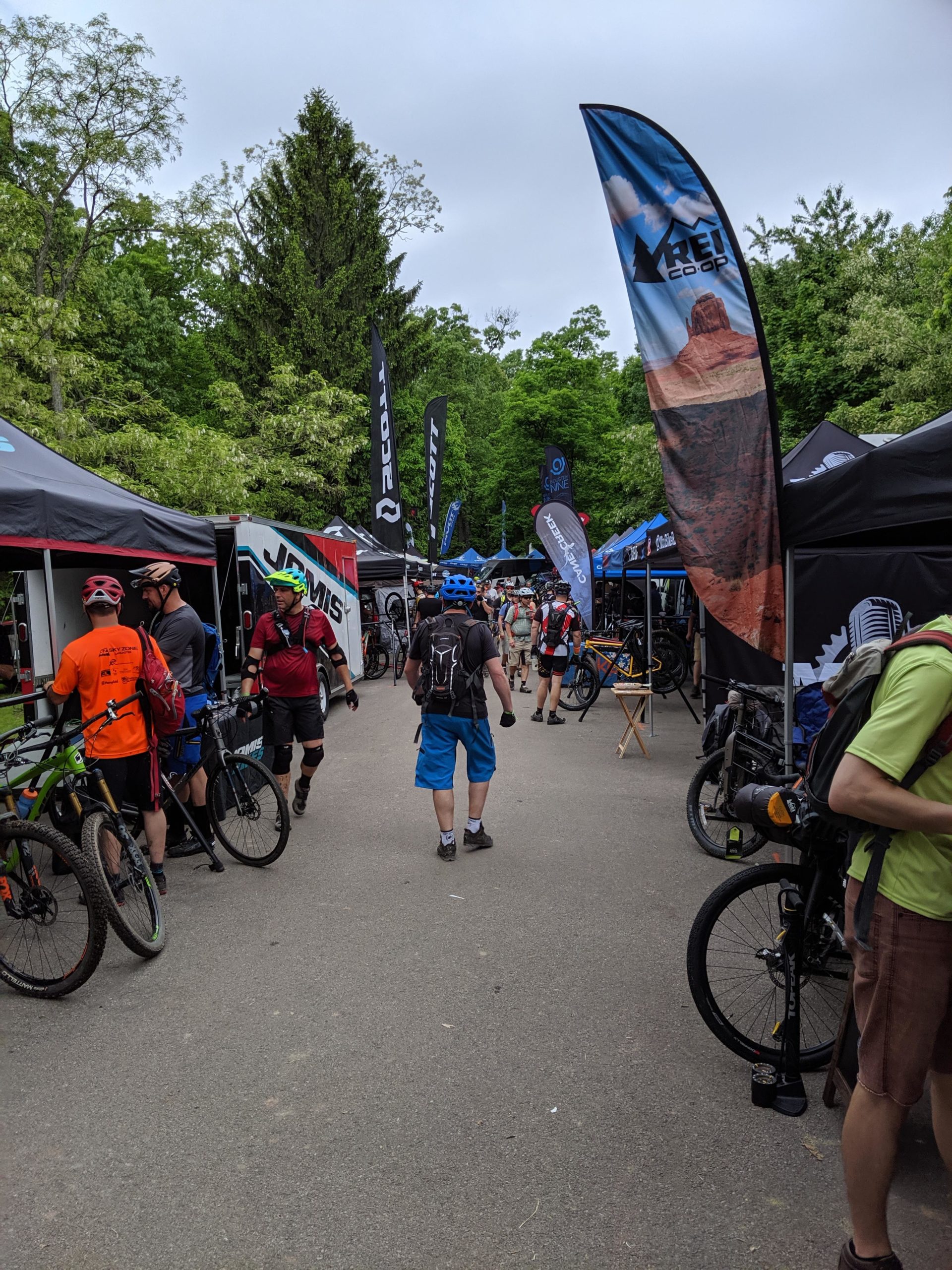 A busy outdoor event with numerous tents and flags from various bicycle brands. Participants in cycling gear are gathered along a pathway lined with bikes, engaging with vendors and exploring the setup. The surrounding area features lush greenery and a cloudy sky, creating a vibrant atmosphere for mountain biking enthusiasts. Allegrippis Trails mountain bike trail.