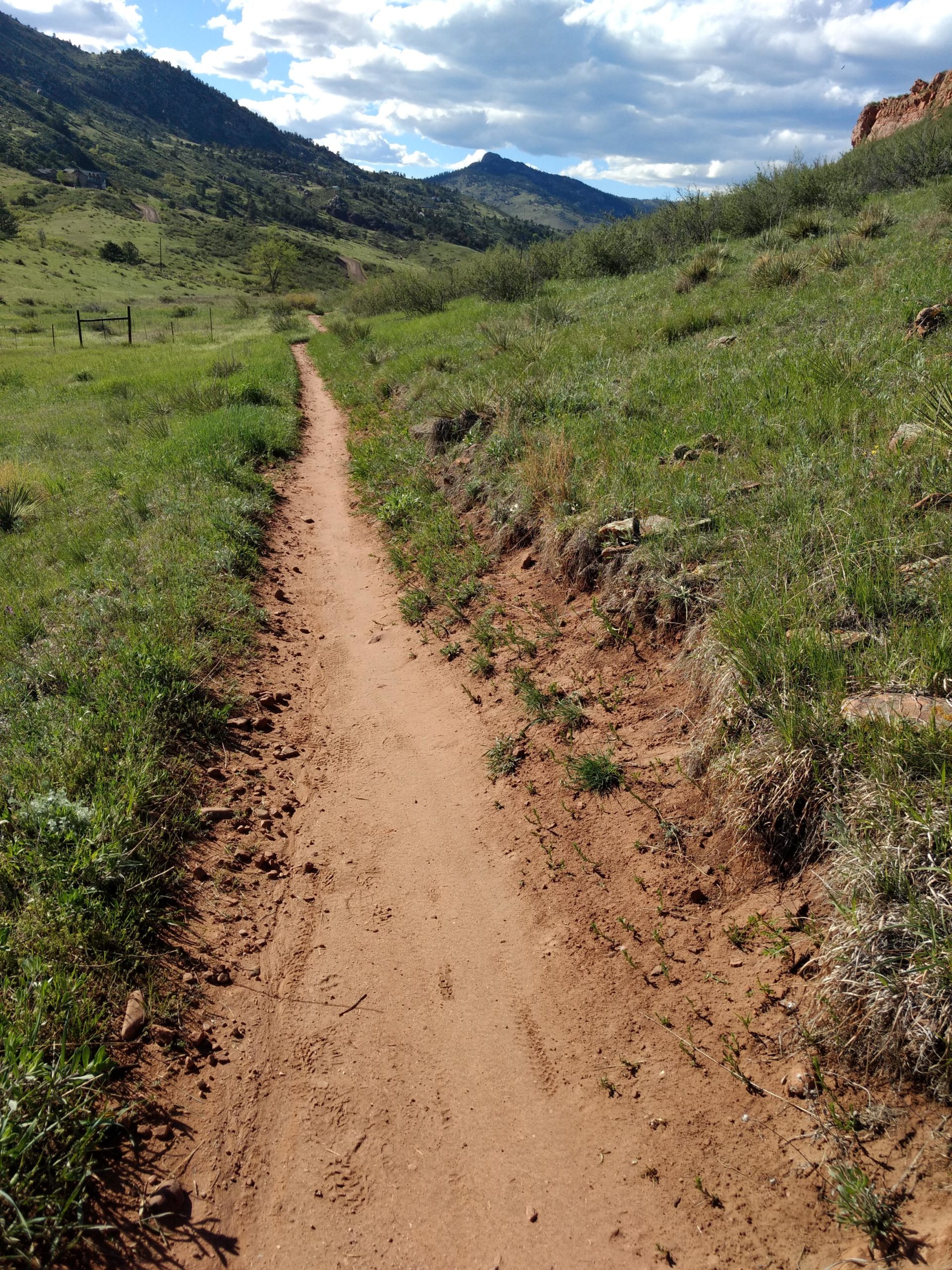 A dirt pathway winds through a green landscape, flanked by sparse grass and small rocks. The trail leads toward distant hills under a partly cloudy sky, with gentle slopes visible on either side. Blue Sky mountain bike trail.