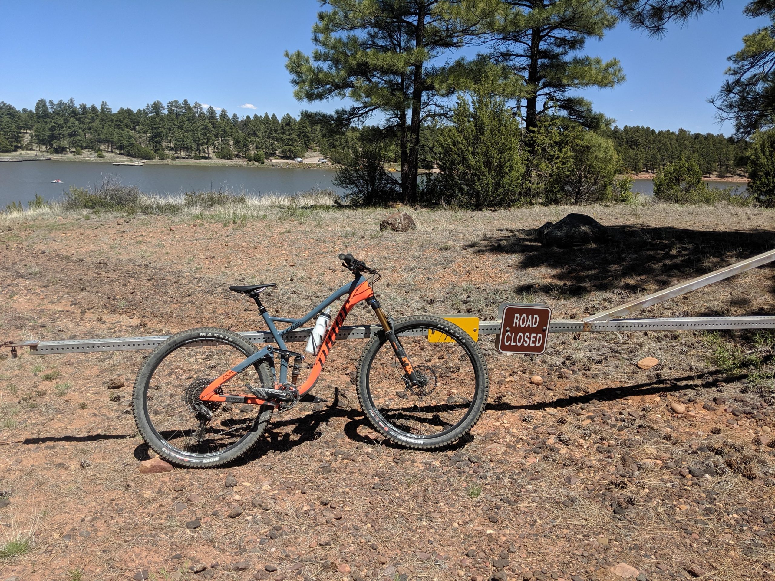 A mountain bike positioned next to a "Road Closed" sign on a gravel path, with a lake and dense pine trees in the background under a clear blue sky. Fools Hollow Lake Loop mountain bike trail.