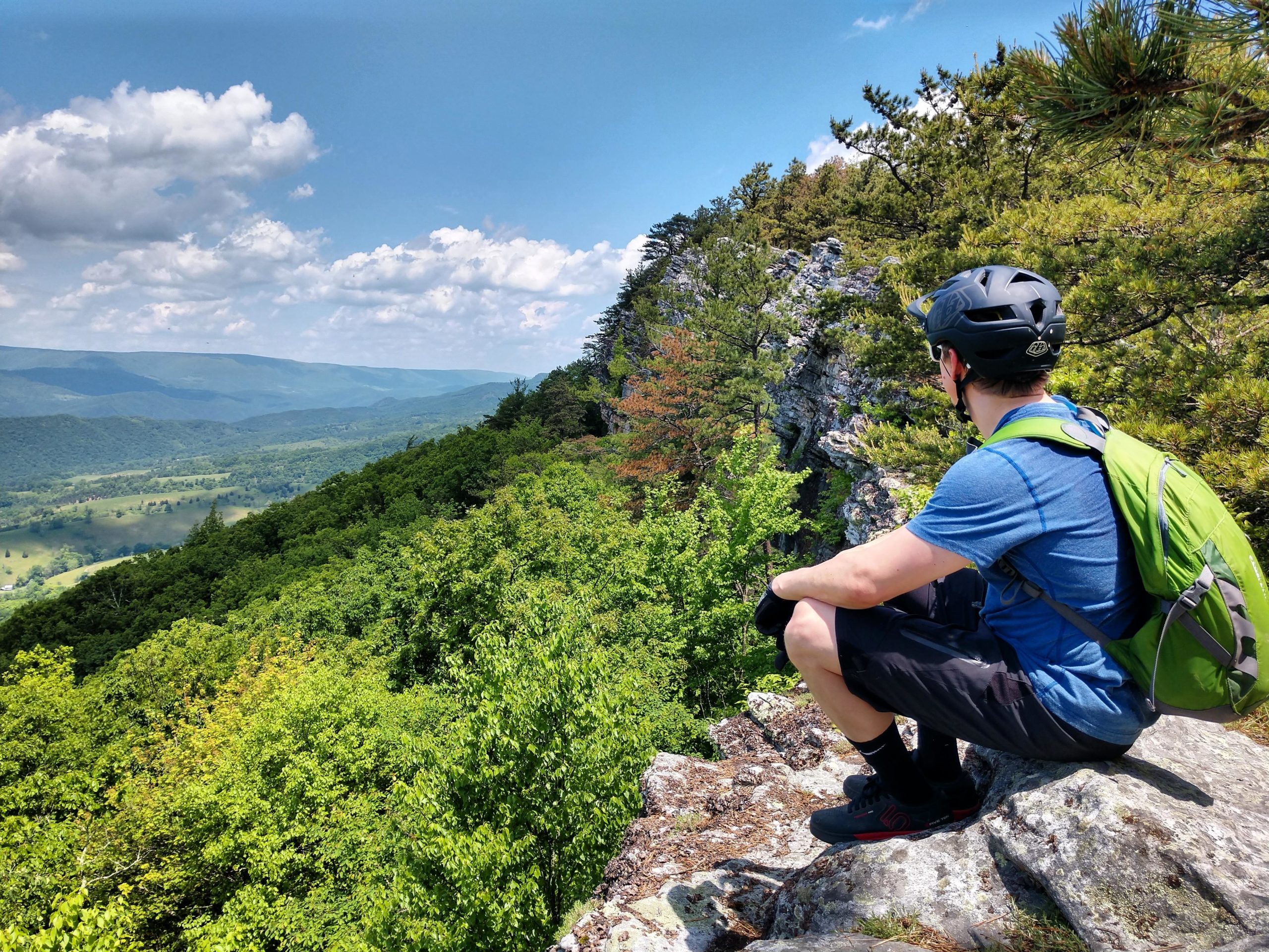 A person sitting on a rocky ledge overlooking a lush green valley and mountains under a partly cloudy sky. The individual is wearing a helmet and a backpack, dressed in casual outdoor attire, enjoying the scenic view of nature. North Fork Mountain Trail mountain bike trail.