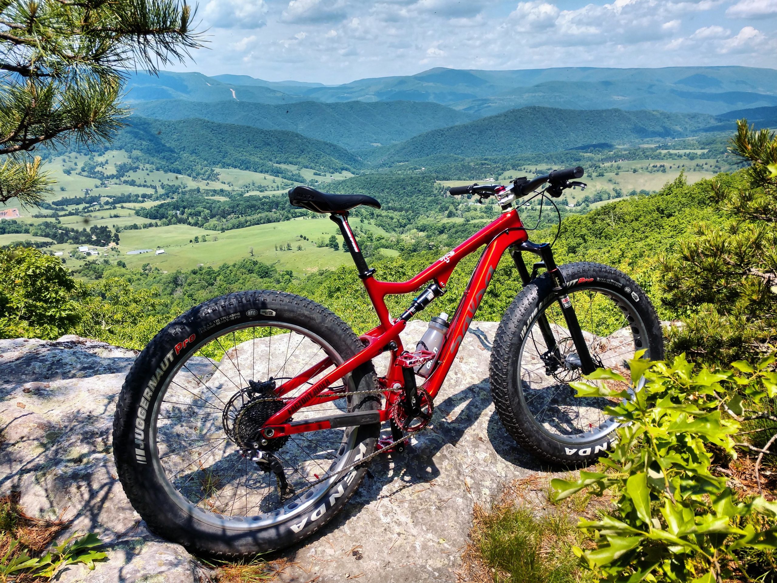 A red mountain bike parked on a rocky outcrop, overlooking a lush green valley and rolling mountains in the background under a partly cloudy sky. North Fork Mountain Trail mountain bike trail.