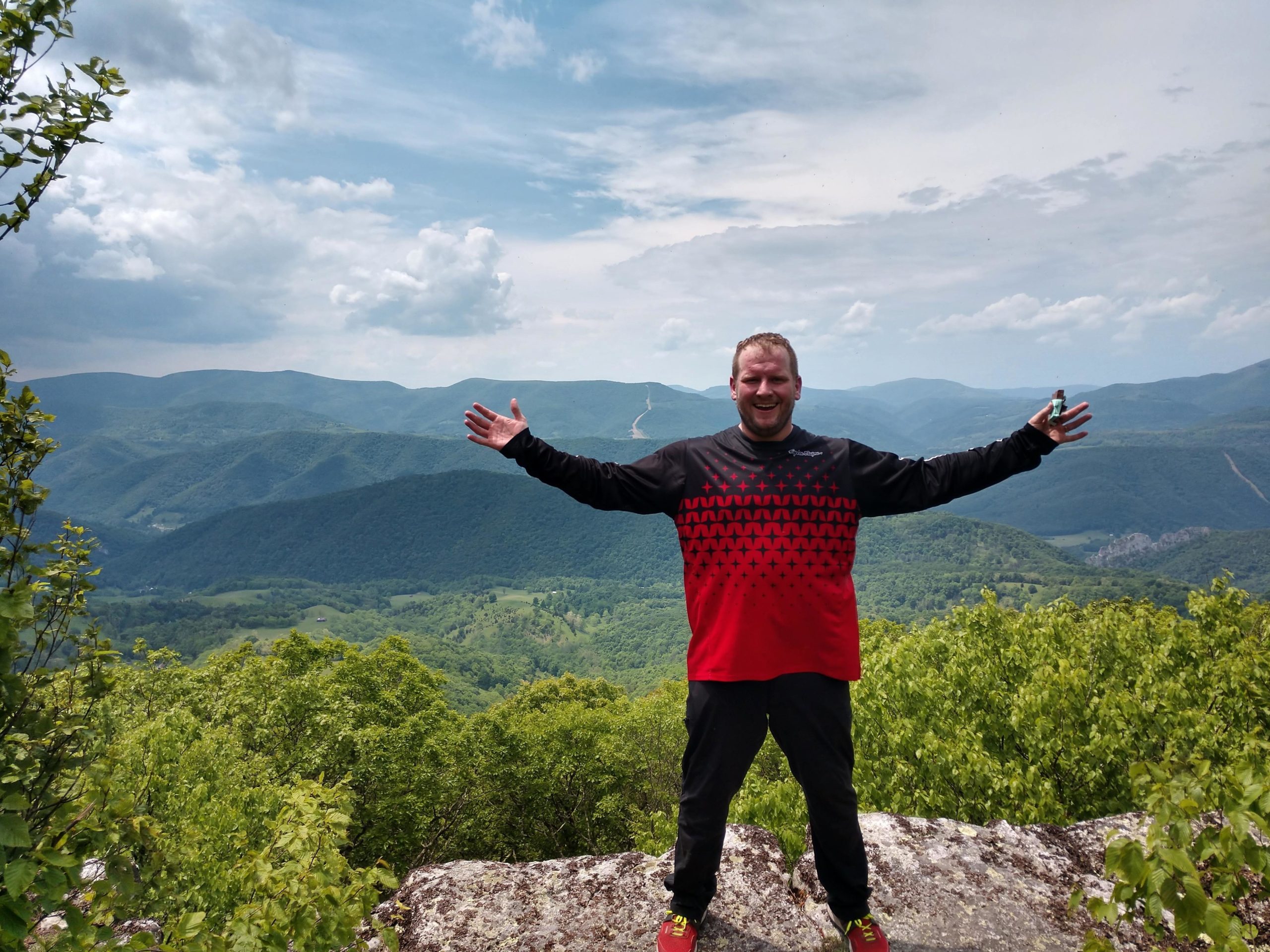 A man stands on a rocky ledge with his arms outstretched, enjoying a scenic view of lush green mountains and a cloudy sky. He is wearing a black and red patterned athletic shirt and black pants. The landscape features rolling hills and a valley below. North Fork Mountain Trail mountain bike trail.