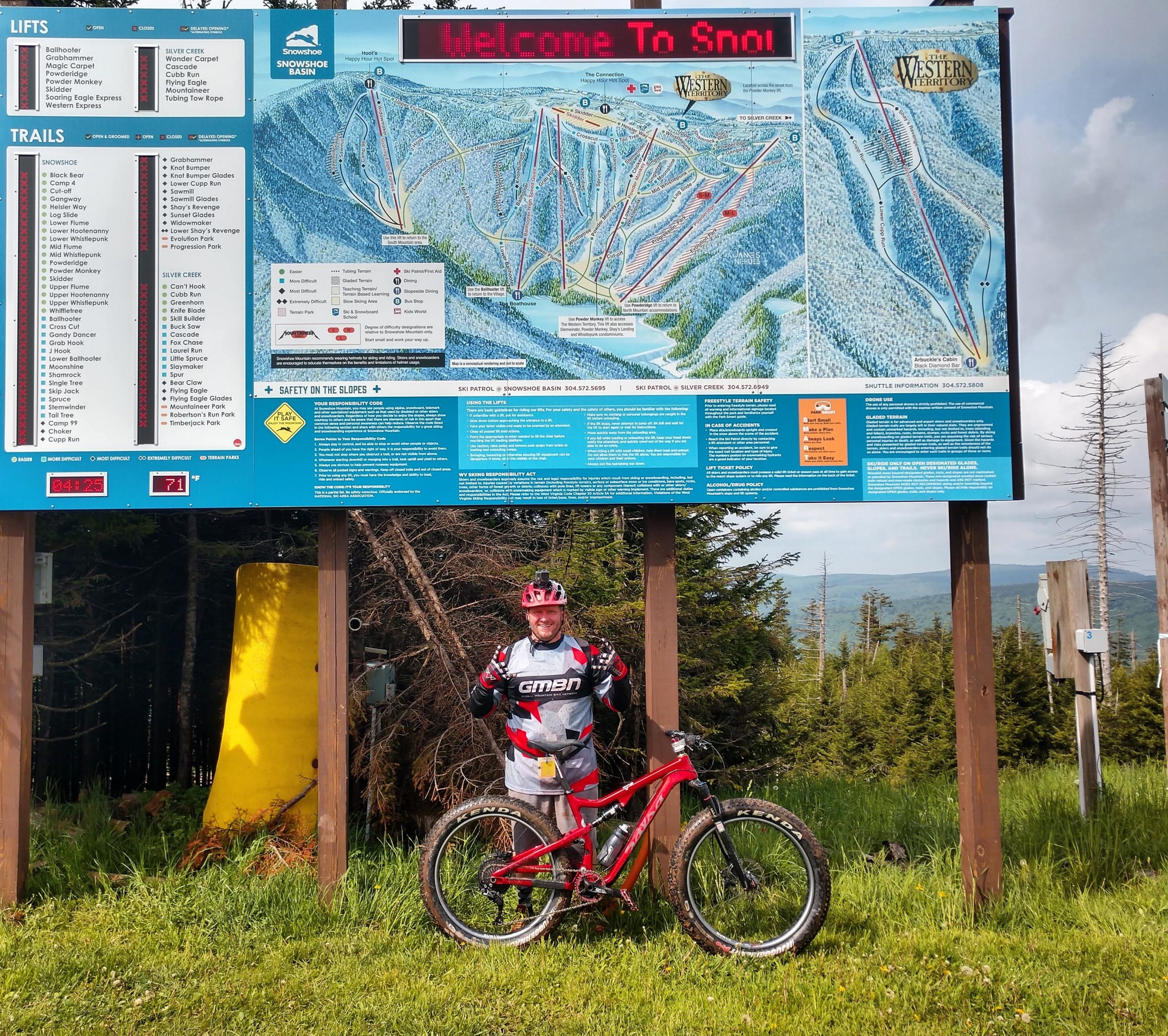 A person in cycling gear stands next to a large informational sign at a bike park, featuring a map of the trails and lifts. The person is smiling and giving a thumbs-up, with a red mountain bike beside them. The background shows trees and a cloudy sky. Snowshoe Bike Park mountain bike trail.