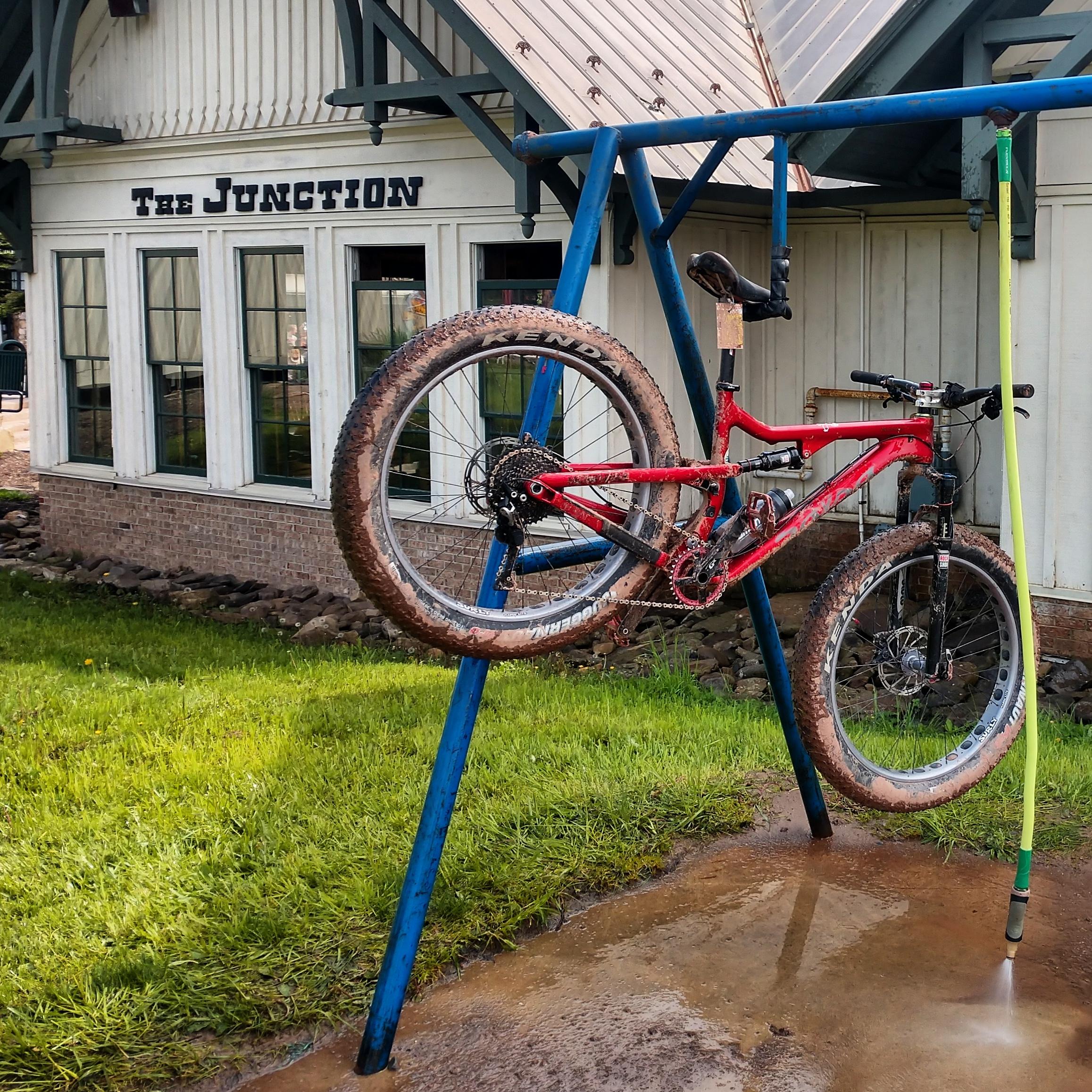 A mountain bike hanging on a blue bike rack for cleaning, with muddy tires. In the background, there is a building with the sign "The Junction." The grass is green, and there is a hose with water spraying on the ground near the bike. Snowshoe Bike Park mountain bike trail.