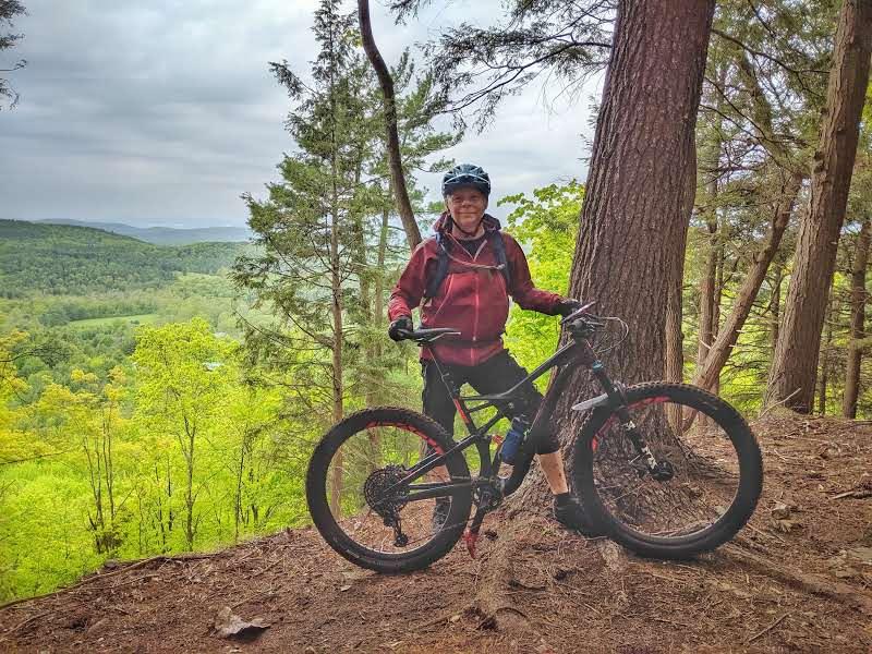 A person wearing a helmet and a red jacket stands next to a mountain bike on a trail, surrounded by trees. In the background, a lush green landscape extends into the distance under a cloudy sky. Fairground Trails mountain bike trail.
