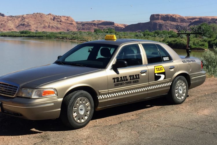 A gold taxi cab parked near a river with cliffs and greenery in the background. The car features a yellow taxi sign on the roof and markings indicating "Trail Taxi" along the side, with additional information about services such as airport transport and bike transport. The scene depicts a clear day, highlighting the natural landscape.