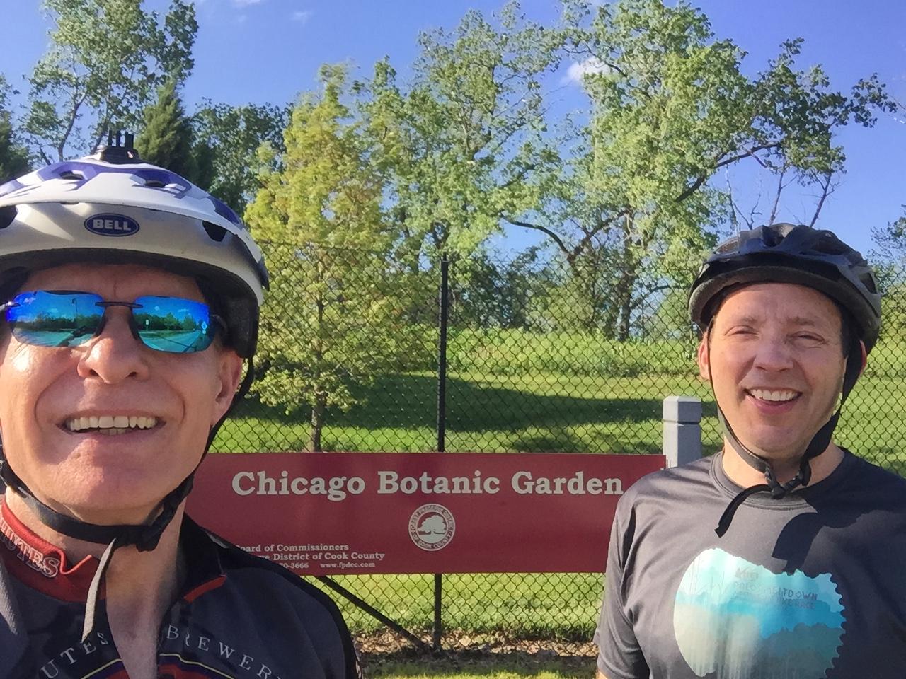 Two men wearing bike helmets and sunglasses smile for a selfie in front of a sign for the Chicago Botanic Garden. They are standing on a sunny day with trees in the background. Caldwell Woods mountain bike trail.