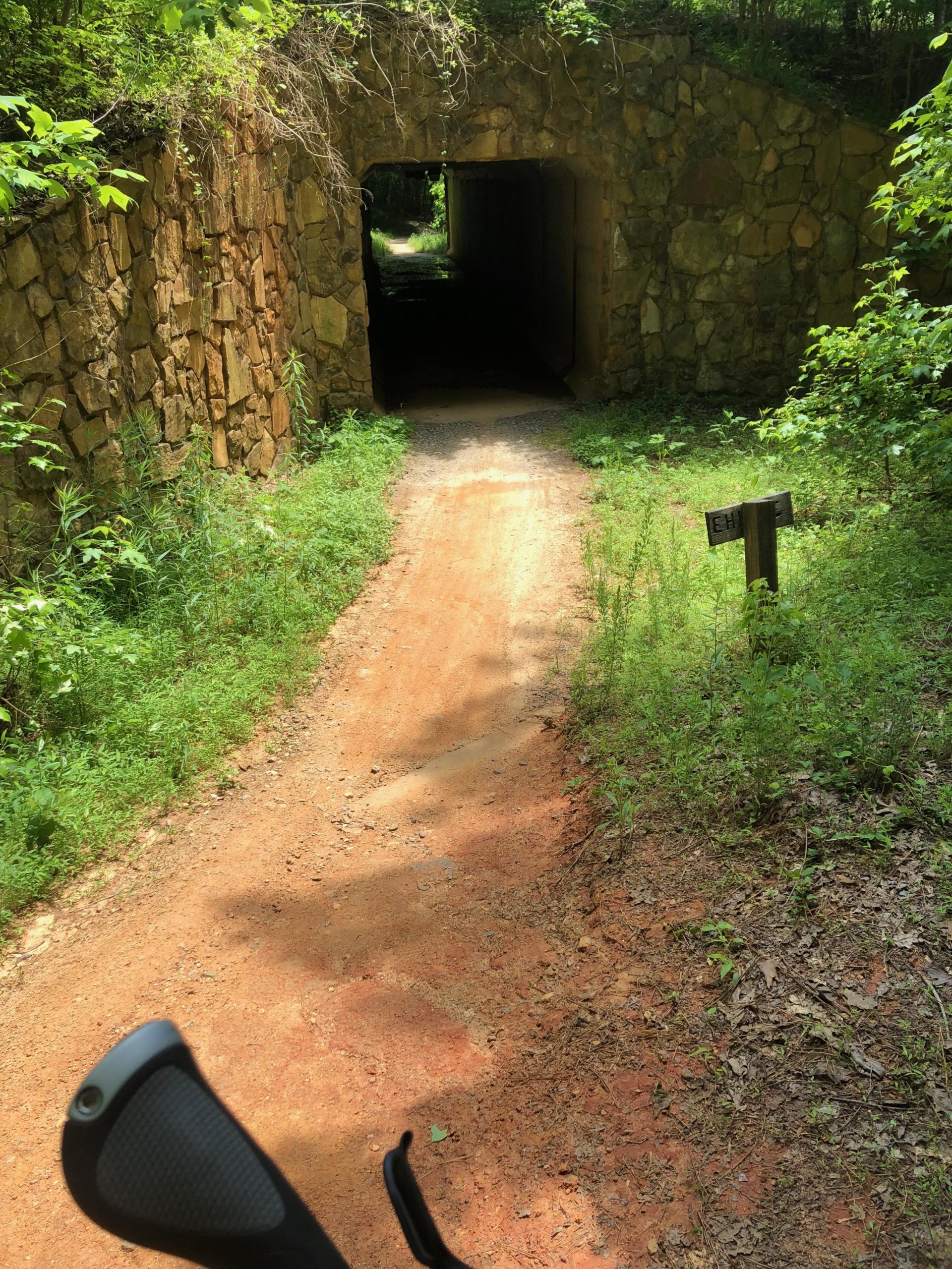 A dirt path leading into a stone tunnel surrounded by lush green foliage, with a wooden sign labeled "EH" to the right. The scene is illuminated by sunlight, highlighting the textures of the earth and vegetation. Anne Springs Close Greenway mountain bike trail.