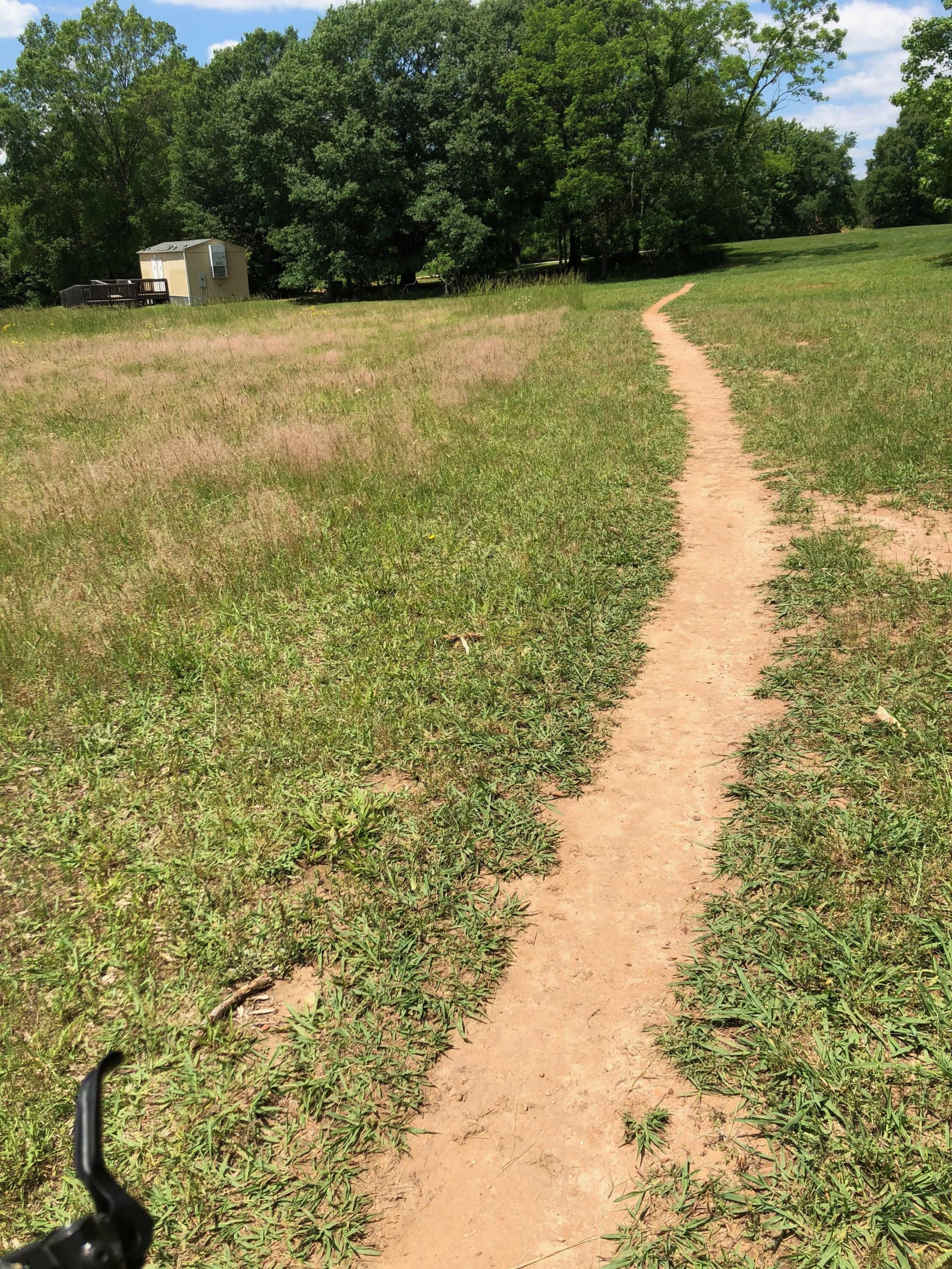 A dirt path winding through a grassy field, bordered by tall green grass and trees. In the background, a small yellow building with a deck is visible, surrounded by a serene natural landscape under a blue sky with scattered clouds. Anne Springs Close Greenway mountain bike trail.