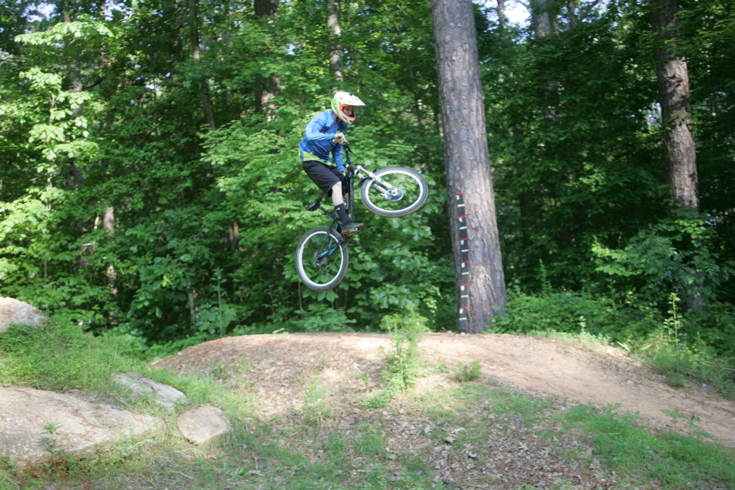 A cyclist in a blue jacket and helmet performs a jump off a dirt ramp in a wooded area, surrounded by lush green trees. The bike is airborne, and the rider's feet are raised off the pedals, showcasing a moment of skillful BMX trick riding. San-lee Park mountain bike trail.