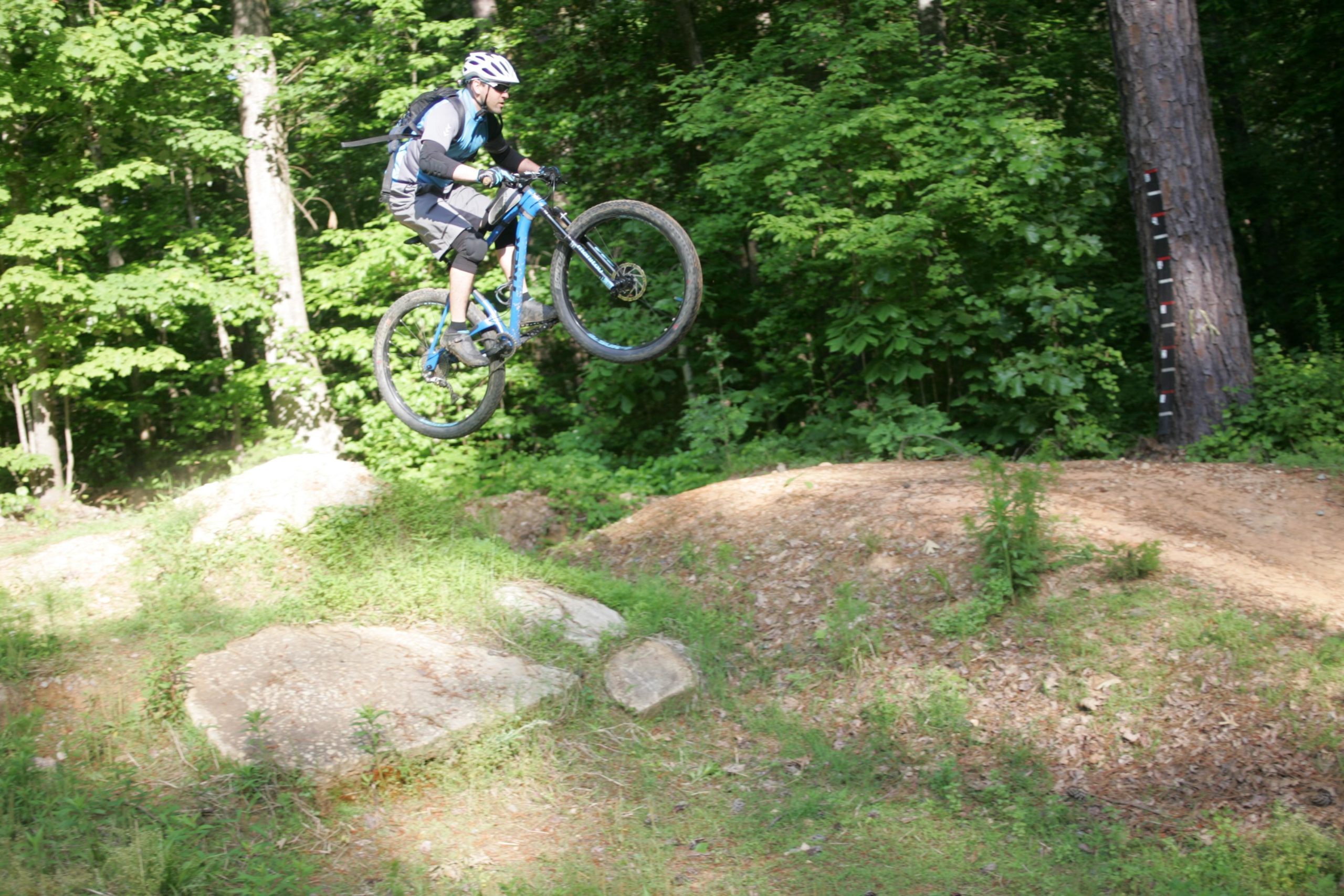 A mountain biker wearing a helmet and protective gear is executing a jump over a dirt mound on a forested trail. Lush green trees surround the scene, providing a natural backdrop. The biker’s blue mountain bike is in mid-air, showcasing the excitement of the sport. San-lee Park mountain bike trail.