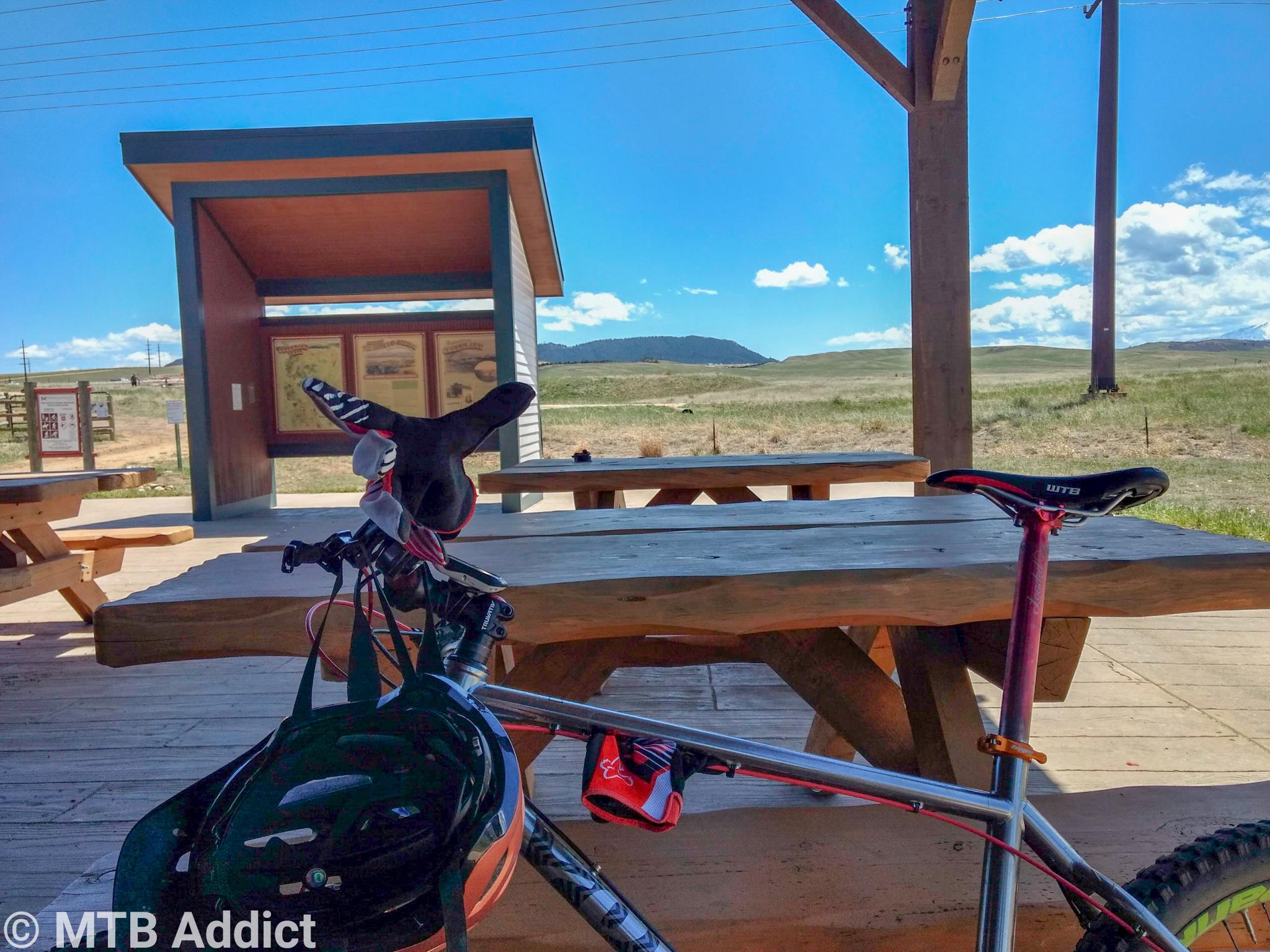 Image of a mountain bike parked at a picnic area with a wooden table, under a shelter. In the background, there is a display board with information and a vast open landscape featuring grass and distant hills under a clear blue sky with a few clouds. A bicycle helmet rests on the bike’s handlebars. Santa Fe Trail mountain bike trail.