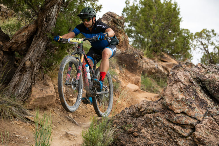 A cyclist in a blue and black jersey and helmet is mid-jump on a rocky mountain biking trail, with trees and rocky terrain in the background. The cyclist is navigating a narrow path surrounded by greenery and large rocks.