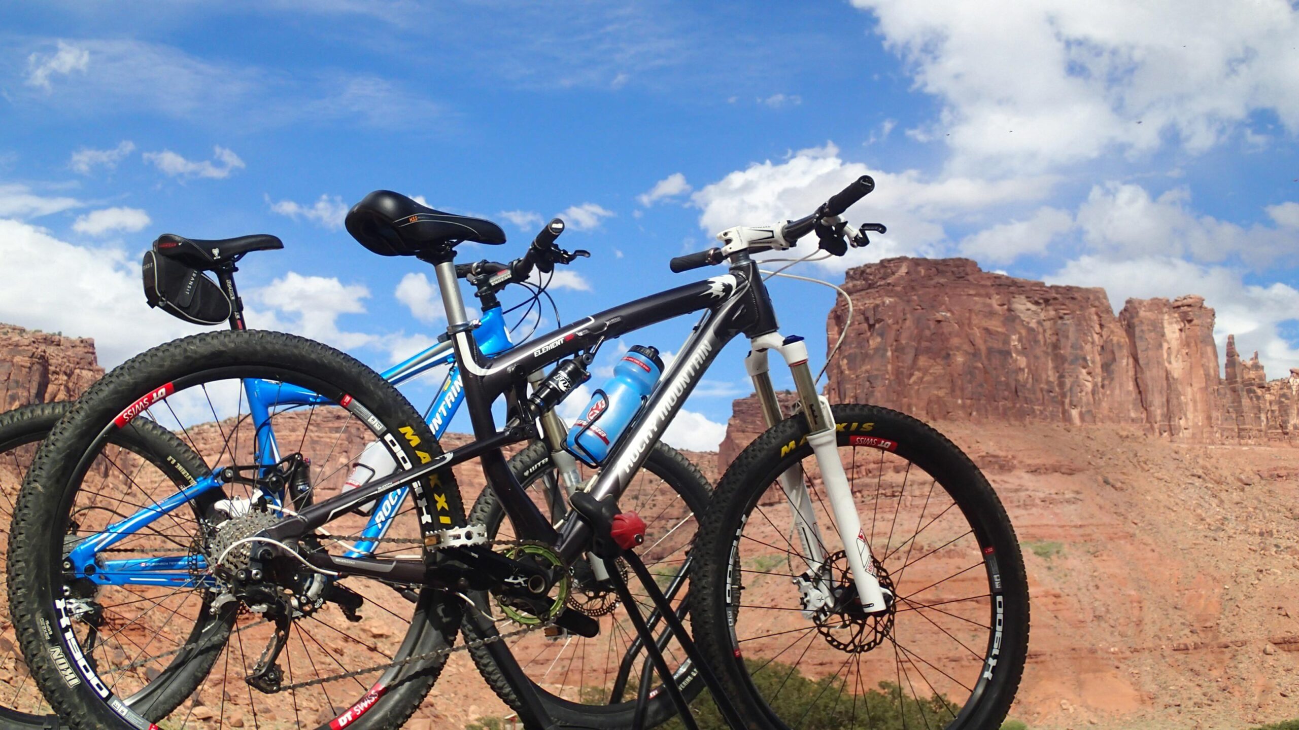 Rocky Mountain Element: Two mountain bikes are parked against a scenic backdrop of red rock formations and a blue sky with fluffy white clouds. The first bike is predominantly black and features a water bottle, while the second bike is bright blue. The landscape suggests a location ideal for outdoor biking adventures.