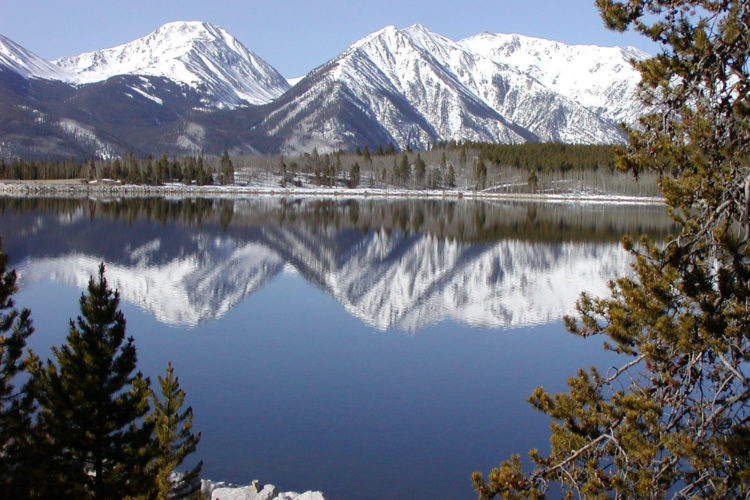 Scenic view of snow-capped mountains reflecting in a calm lake, framed by evergreen trees along the shoreline.