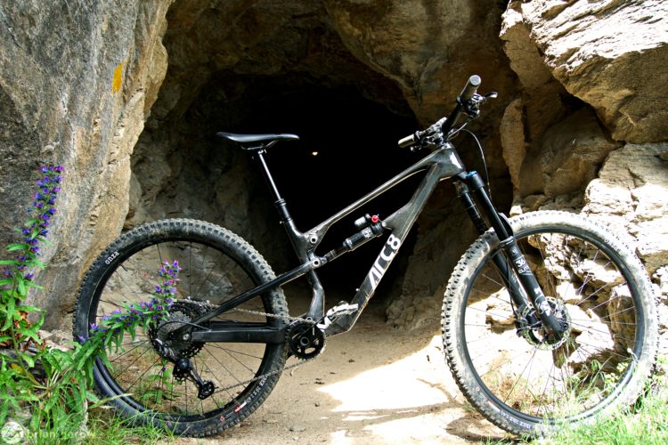 A black mountain bike with a sleek design is positioned next to a rocky entrance of a cave. Vibrant purple flowers and green foliage are visible in the foreground, adding a natural touch to the outdoor setting. The cave looms in the background, suggesting an adventurous atmosphere.