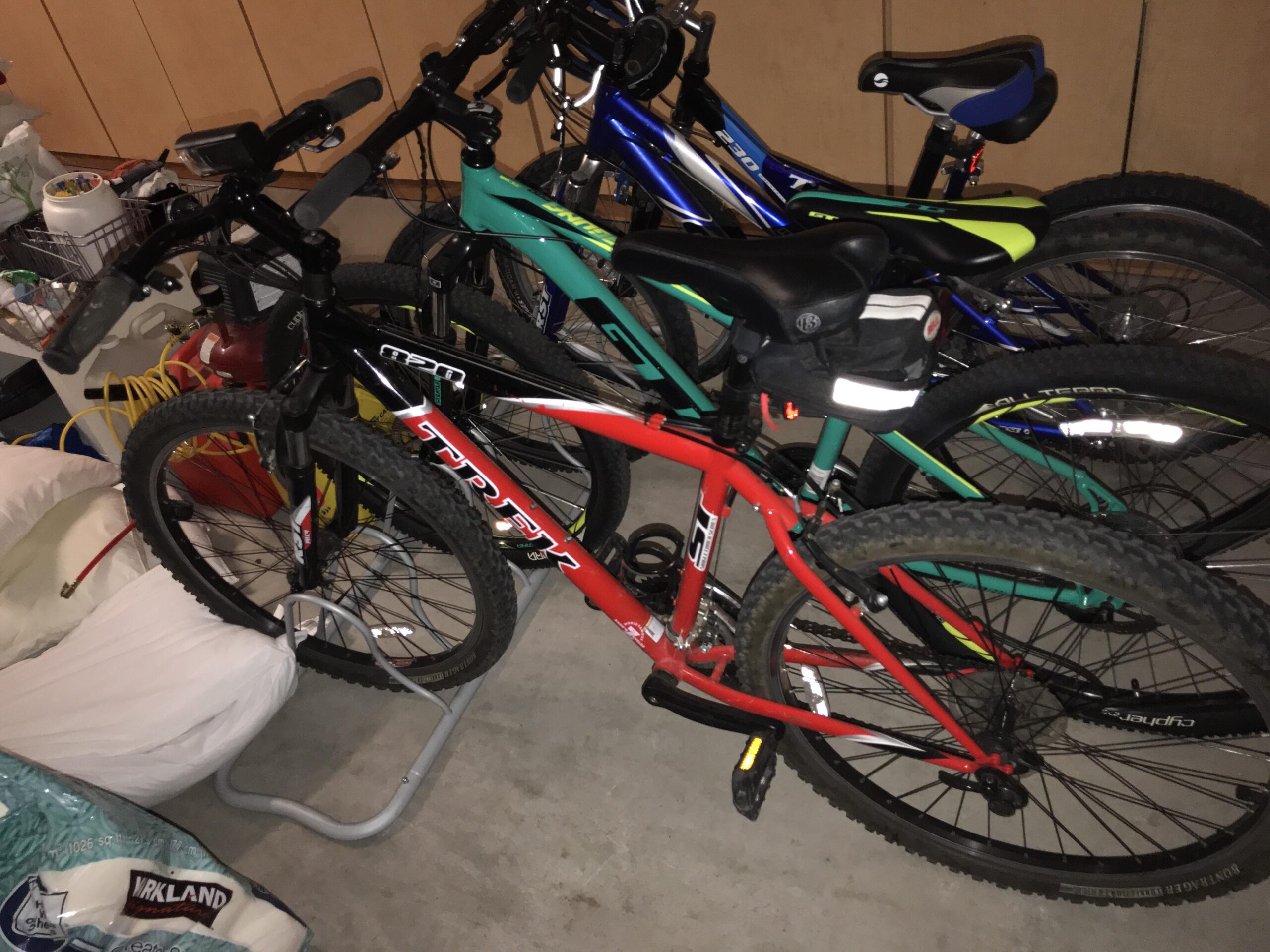 Trek 820: A cluttered garage with several bicycles parked closely together. The foreground features a red mountain bike with thick tires, while several other bikes in various colors, including blue and green, are visible in the background. Various items, such as bags and tools, are scattered around on the floor, adding to the packed appearance of the space.