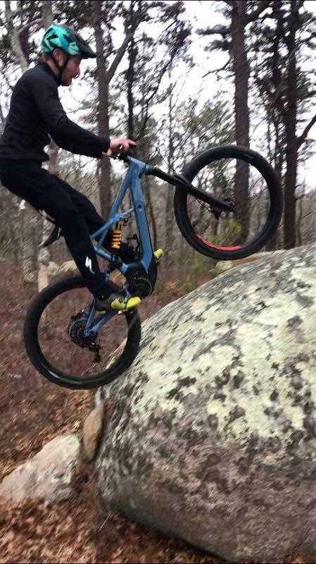 A mountain biker in a black long-sleeve shirt and pants performs a jump off a large boulder in a forested area. The biker is mid-air, with one wheel raised above the rock, surrounded by trees and fallen leaves on the ground. Otis mountain bike trail.