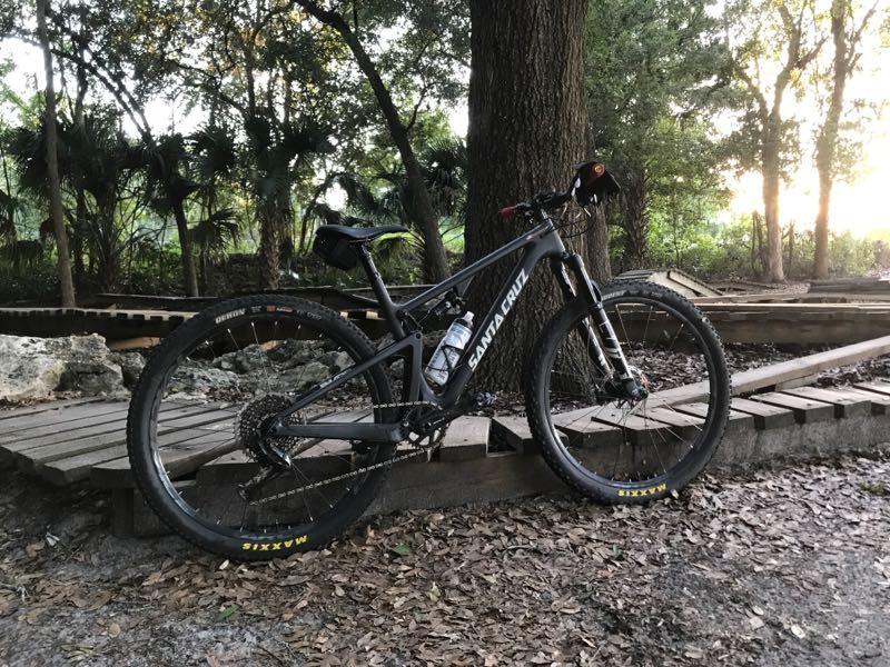 A mountain bike leaning against a tree, positioned on a wooden pathway surrounded by greenery. The bike has a sleek black frame with the brand name "Santa Cruz" prominently displayed. Soft sunlight filters through the trees, creating a serene outdoor setting. Mount Dora Trail mountain bike trail.